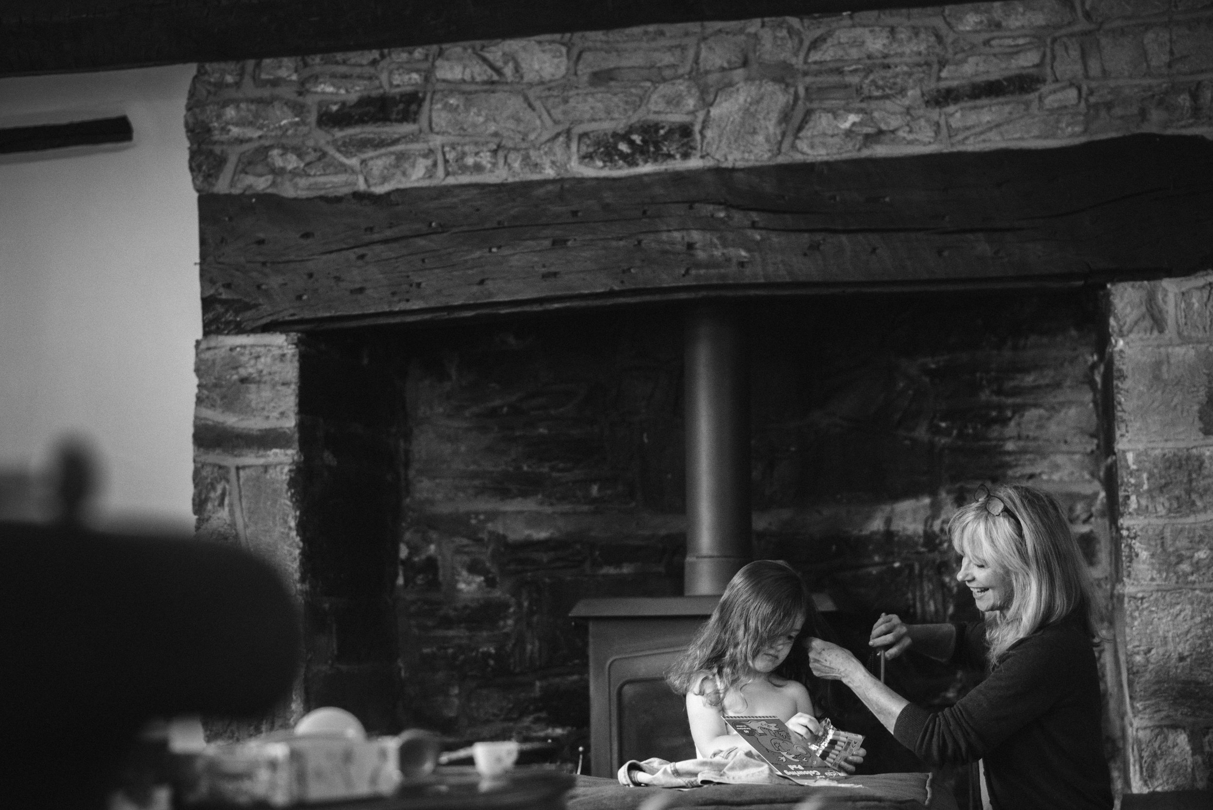 A woman with long blonde hair and glasses smiling as she puts makeup on a young girl sitting at a table in front of a stone fireplace.
