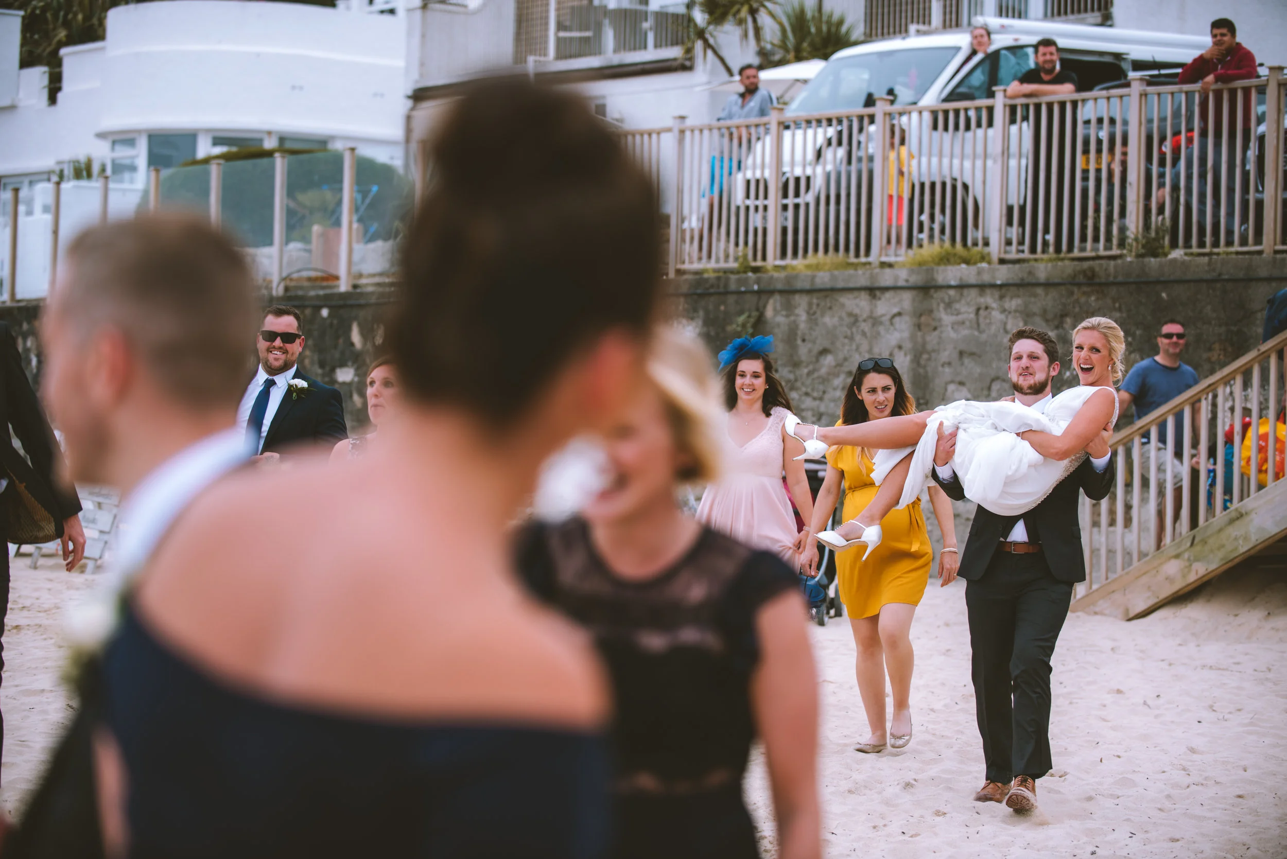 A man in a suit carrying a woman in a white dress on the beach at a wedding celebration, with guests laughing and watching.