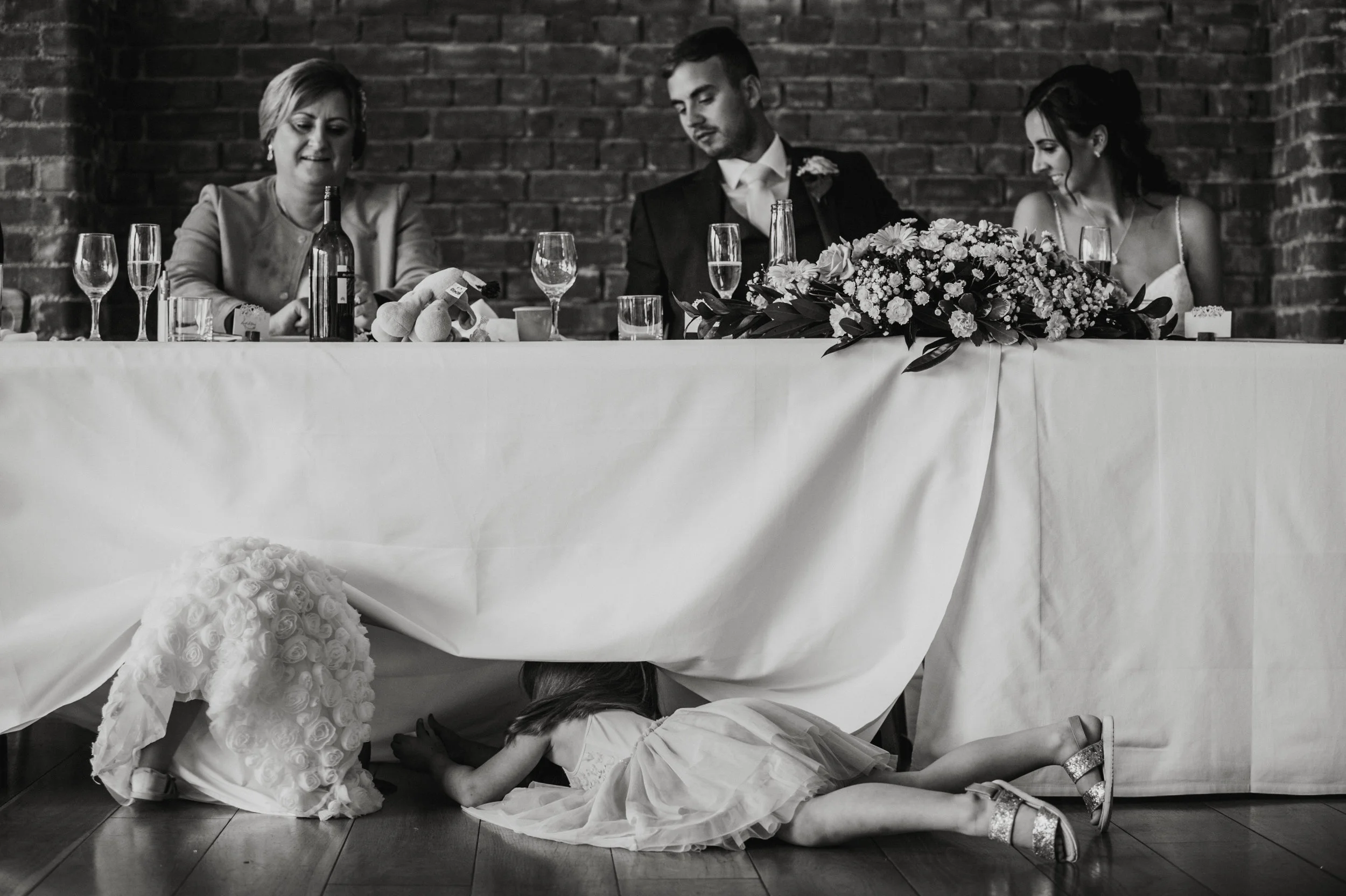 Two young girls are crawling under a decorated table at a wedding reception, with a bride and groom sitting at the table behind them. The table has a white cloth, floral centerpiece, and several glasses and bottles. The bride is dressed in white and 