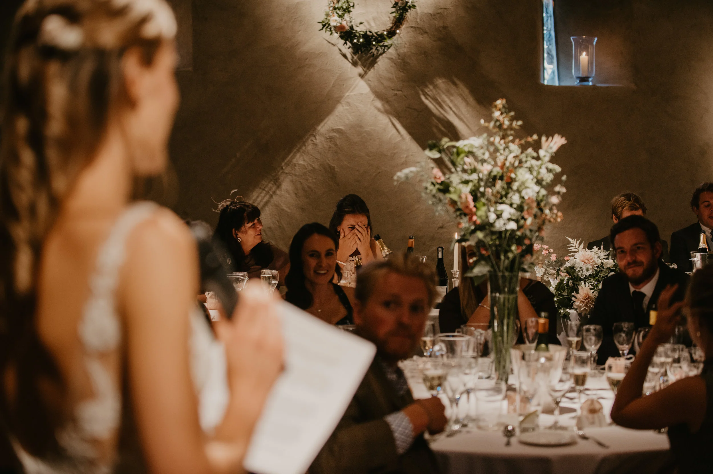 Wedding reception with guests seated at a decorated table, woman giving a speech, flowers, and candle-lit window.