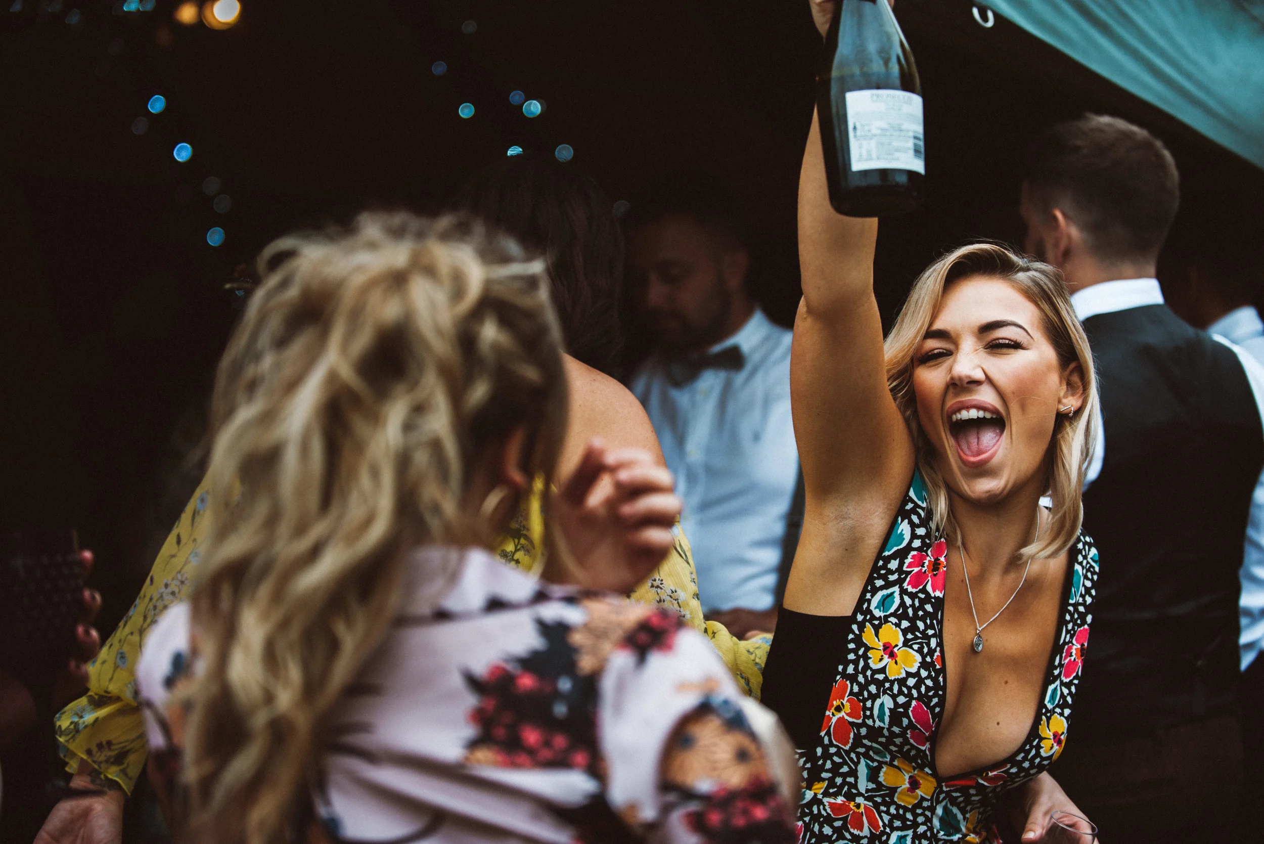 A woman with blonde hair celebrating with an excited expression, holding a champagne bottle in the air at a party or celebration.