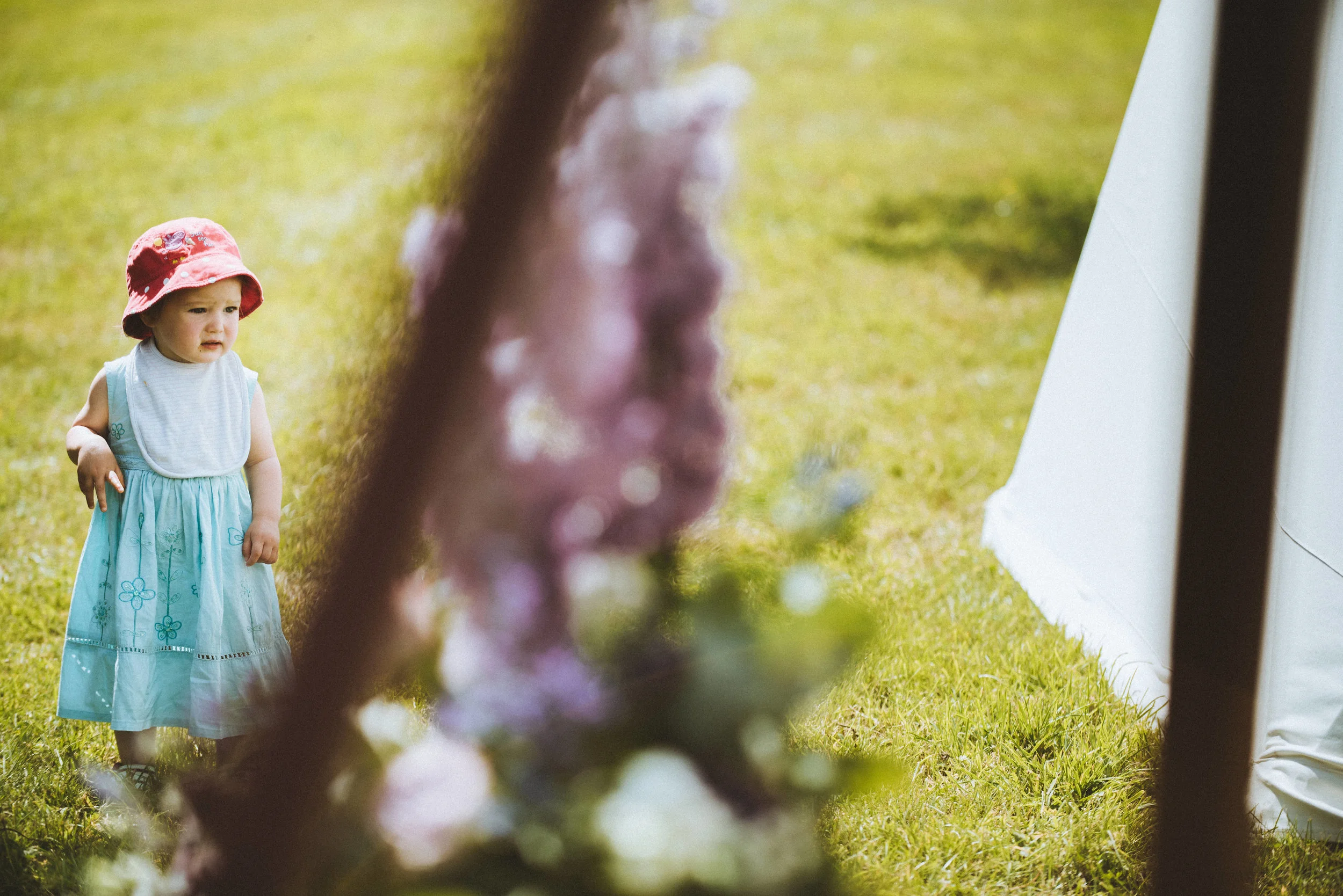 Wedding captured by Cornwall wedding photographer Mark Shaw Photography - A young girl in a blue dress and pink hat standing outside on grass, looking at something with a curious expression.
