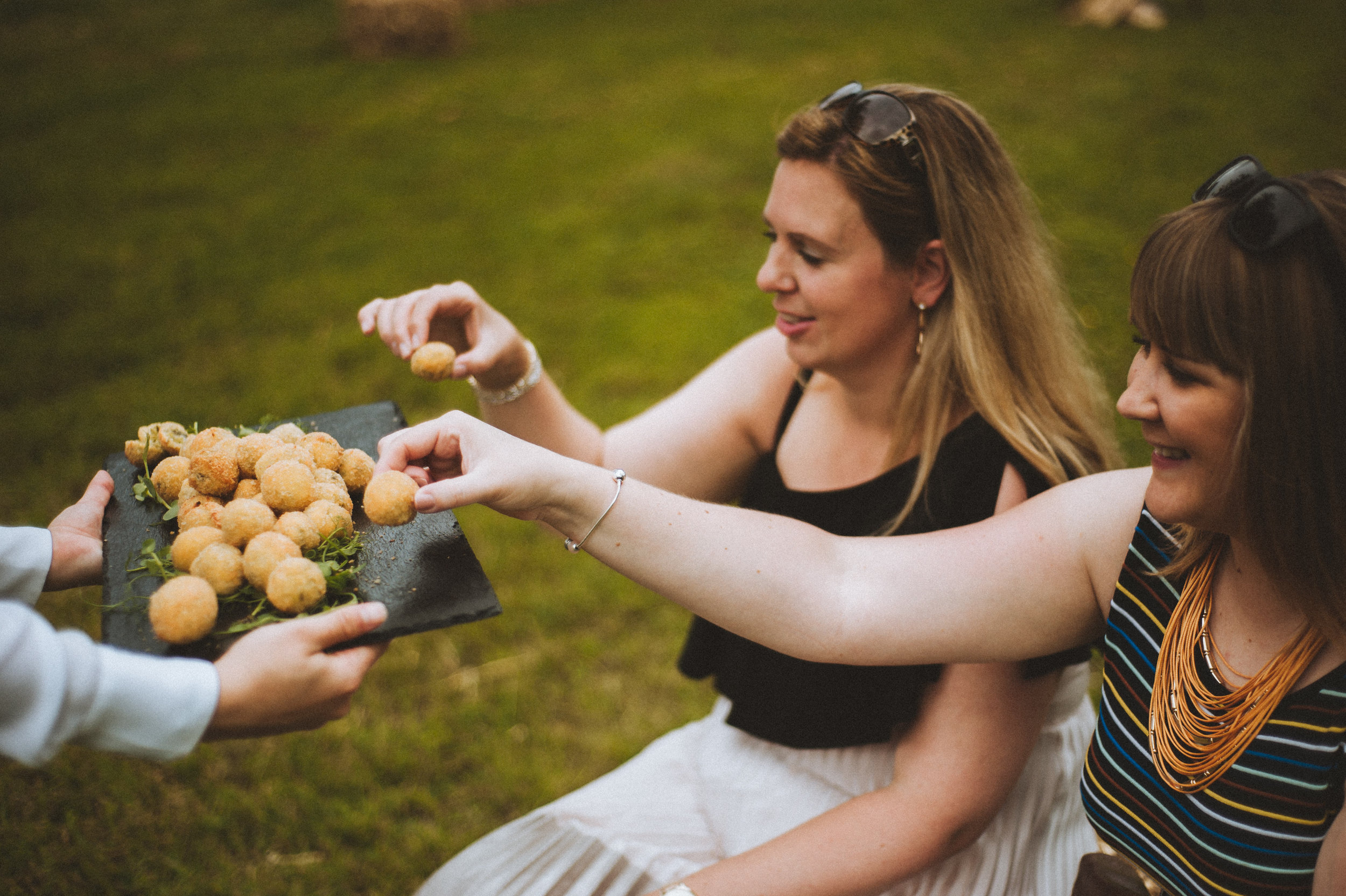 Wedding captured by Cornwall wedding photographer Mark Shaw Photography - Two women are smiling and reaching for a plate of fried food handed by someone off camera, outdoors on grass.