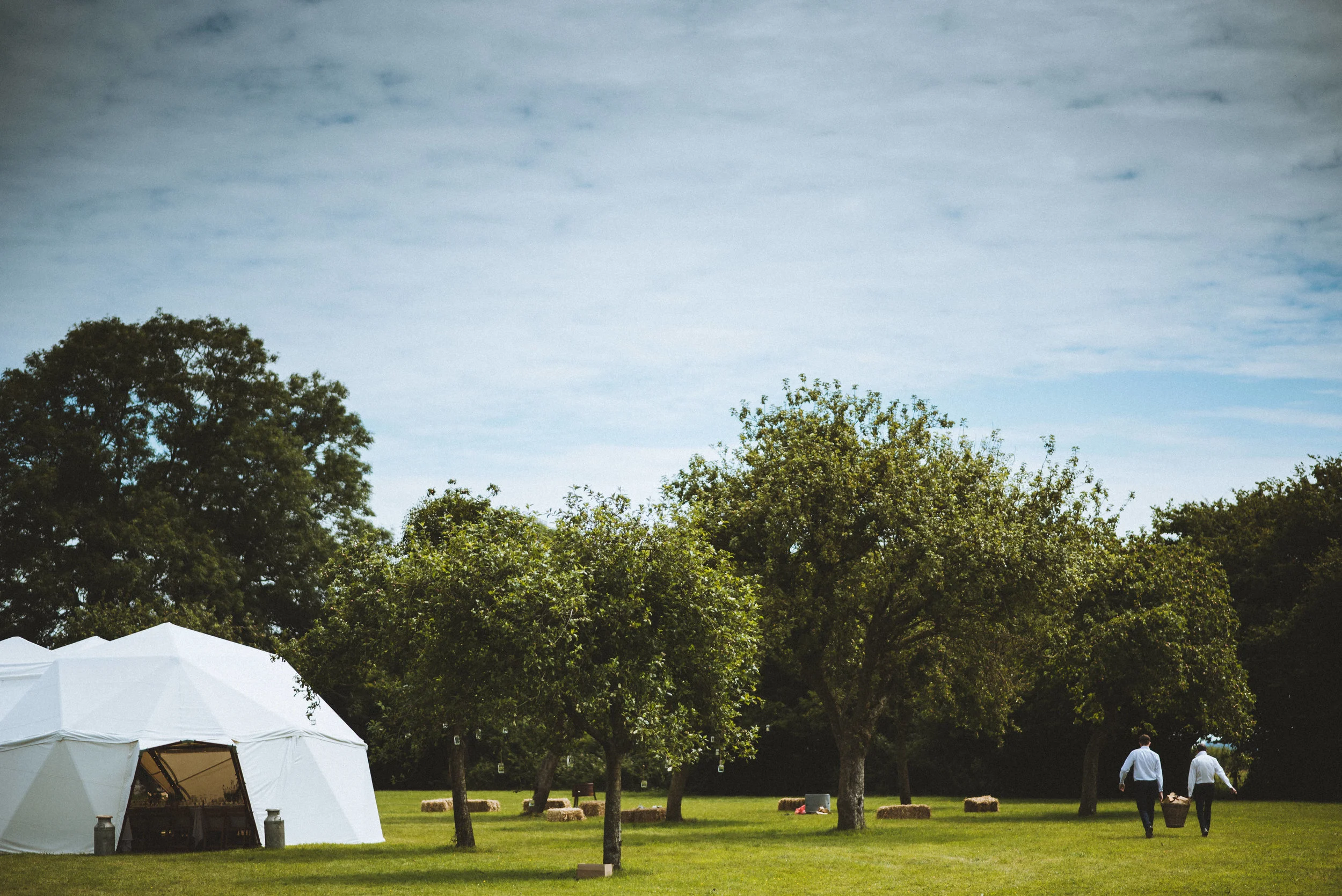 Pengenna manor wedding photography captured by Cornwall wedding photographer Mark Shaw Photography -A park with green grass, trees, a white event tent, and two people walking with a basket.
