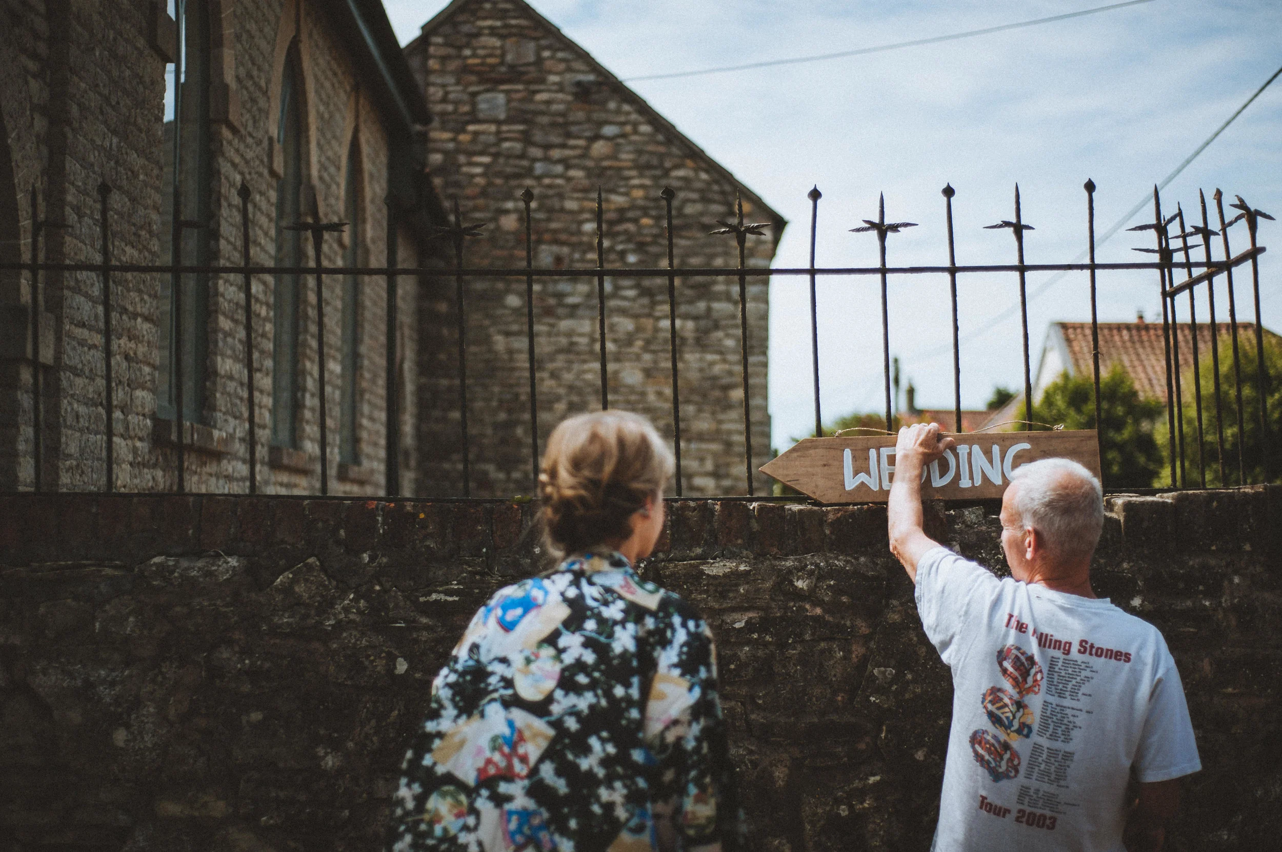 Pengenna manor wedding photography captured by Cornwall wedding photographer Mark Shaw PAn older man in a white T-shirt installing a wooden wedding sign on a stone wall, with a woman standing nearby, against a backdrop of a stone building and houses.