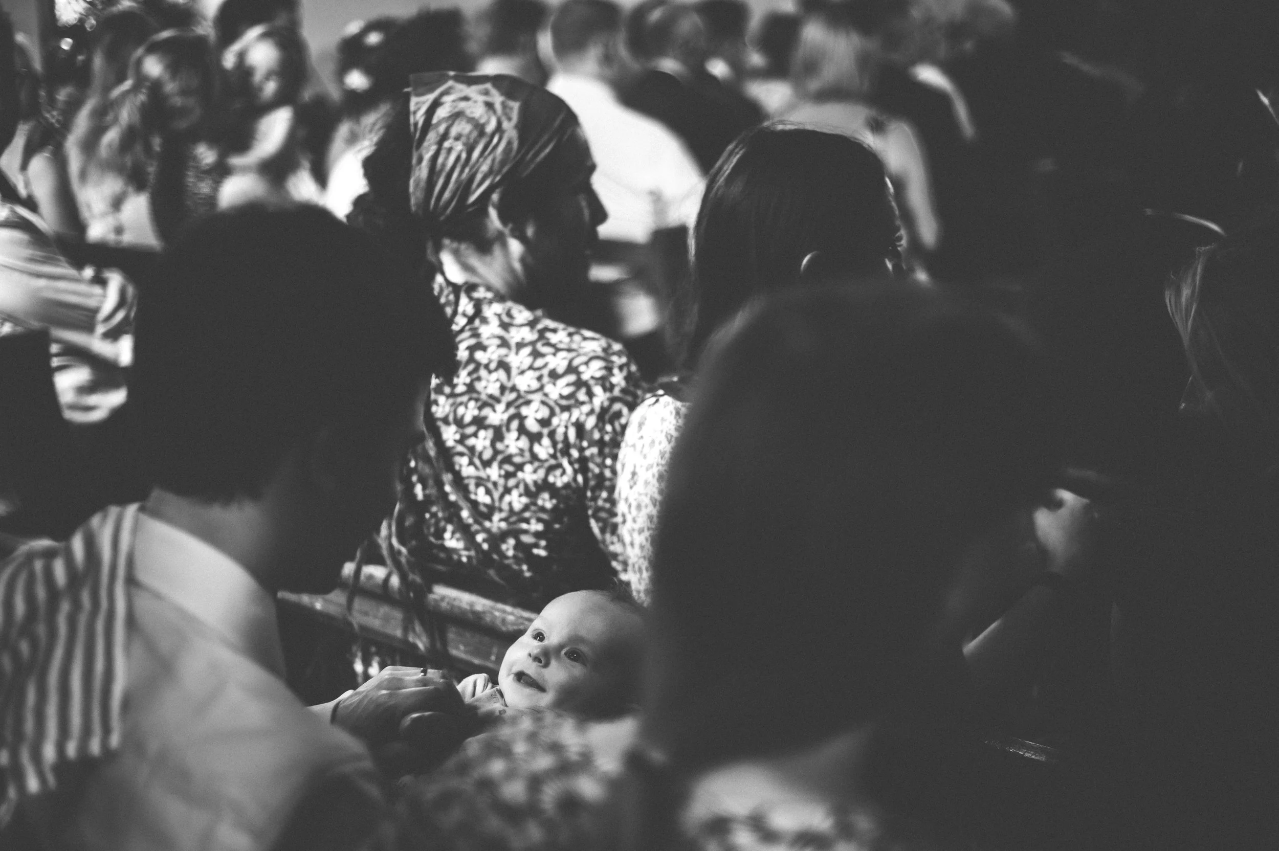 Wedding captured by Cornwall wedding photographer Mark Shaw Photography - A black and white photo of a crowded audience with various people sitting, including a woman with a patterned headscarf and a baby looking up at her, in an indoor setting.