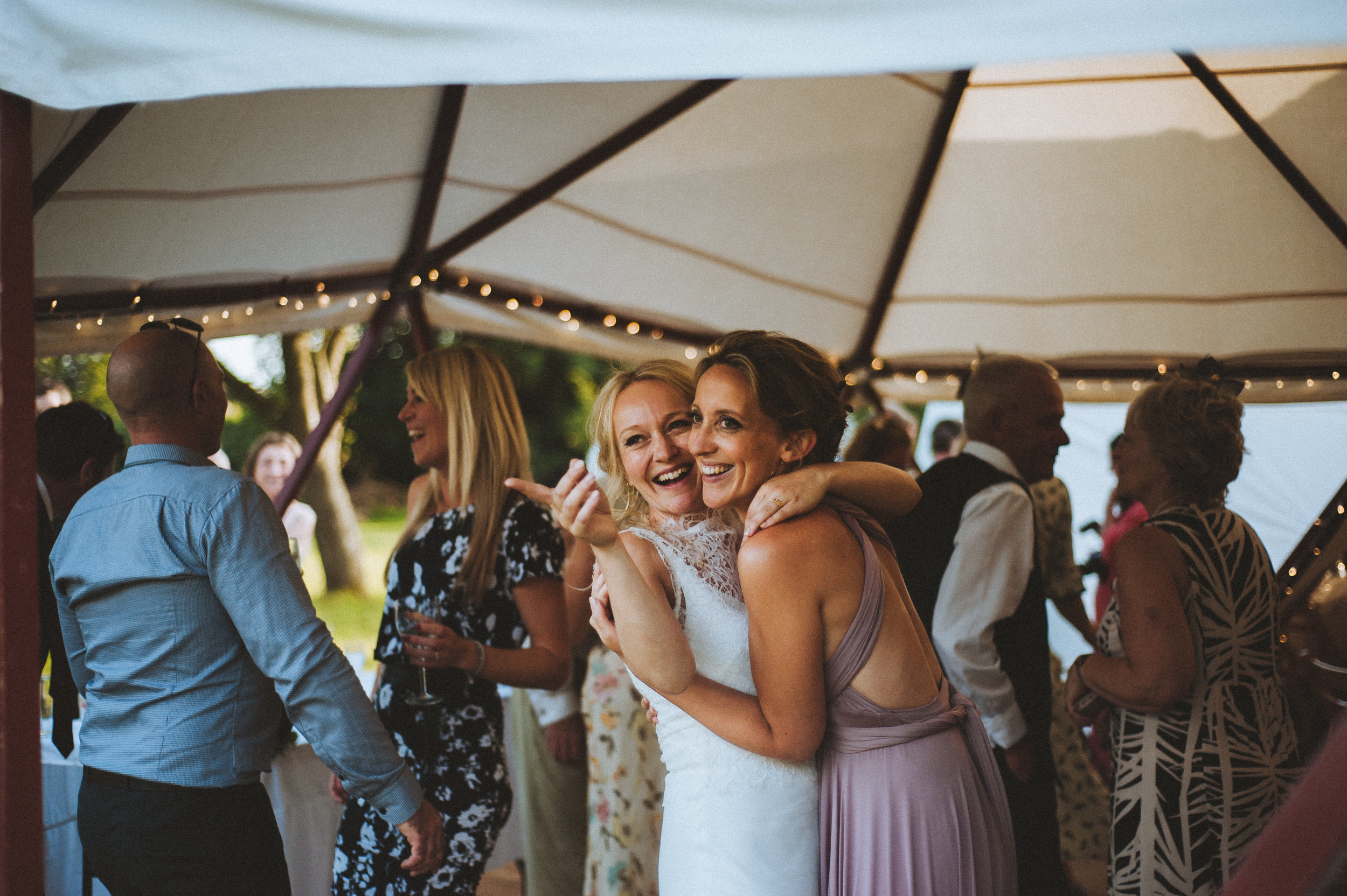 Wedding captured by Cornwall wedding photographer Mark Shaw Photography - Two women hugging and smiling at a party under a tent, with other guests dancing and talking in the background.