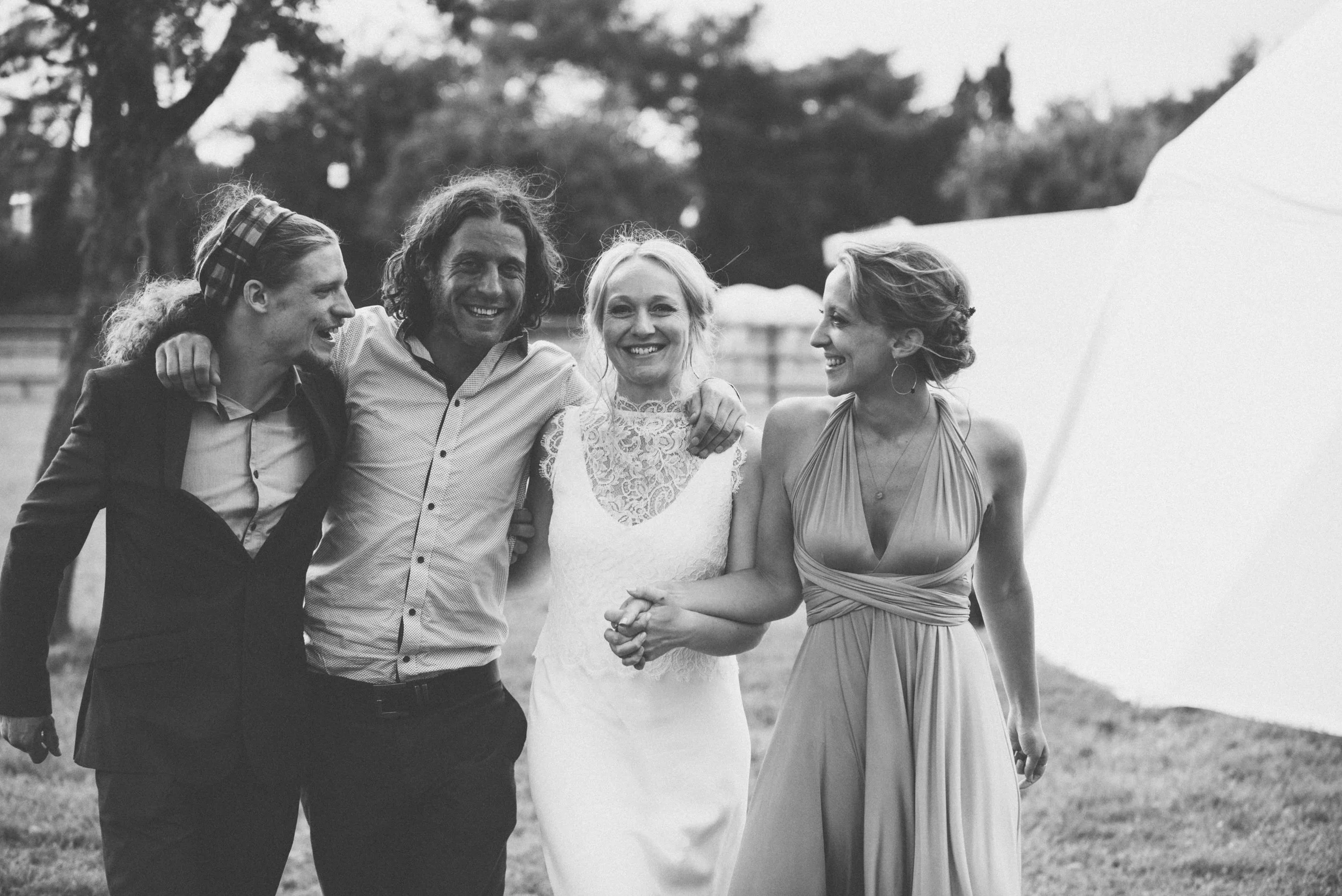 Wedding captured by Cornwall wedding photographer Mark Shaw Photography - Black and white photo of four people smiling and walking arm in arm outdoors, near a large tent with trees in the background. 