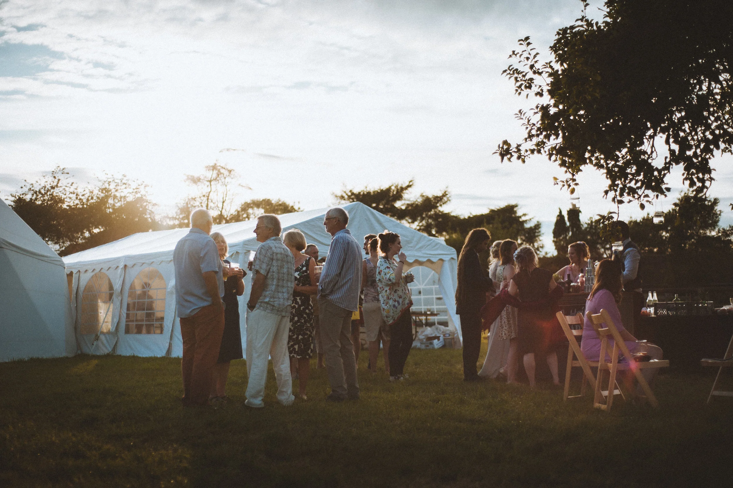 Wedding captured by Cornwall wedding photographer Mark Shaw Photography - People gathered outdoors in the evening near a white tent, socializing and enjoying drinks, with trees and a cloudy sky in the background.