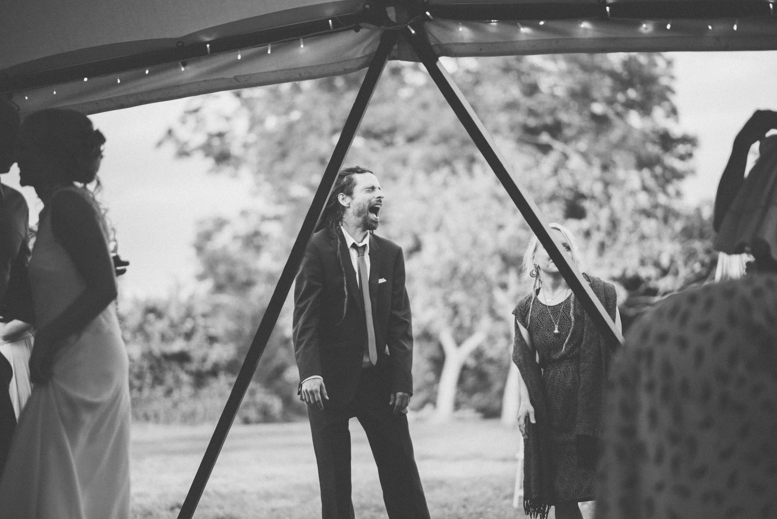 A black and white photo of a man in a suit, laughing with his mouth open, standing outdoors under a canopy at a gathering. Several people are around him, some partially visible, with trees in the background.