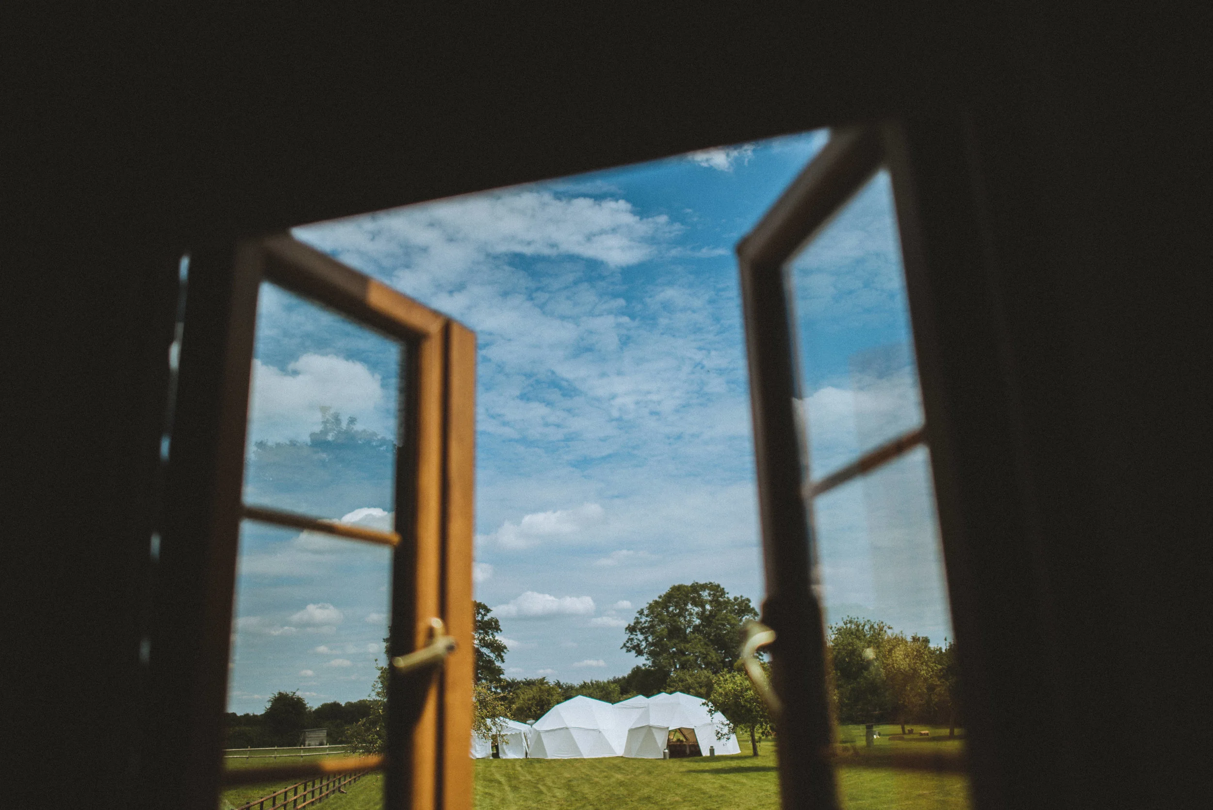 Pengenna manor wedding photography captured by Cornwall wedding photographer Mark Shaw Photography - Looking out of a window with open wooden shutters, showing a green field with trees, and white structures under a partly cloudy blue sky.