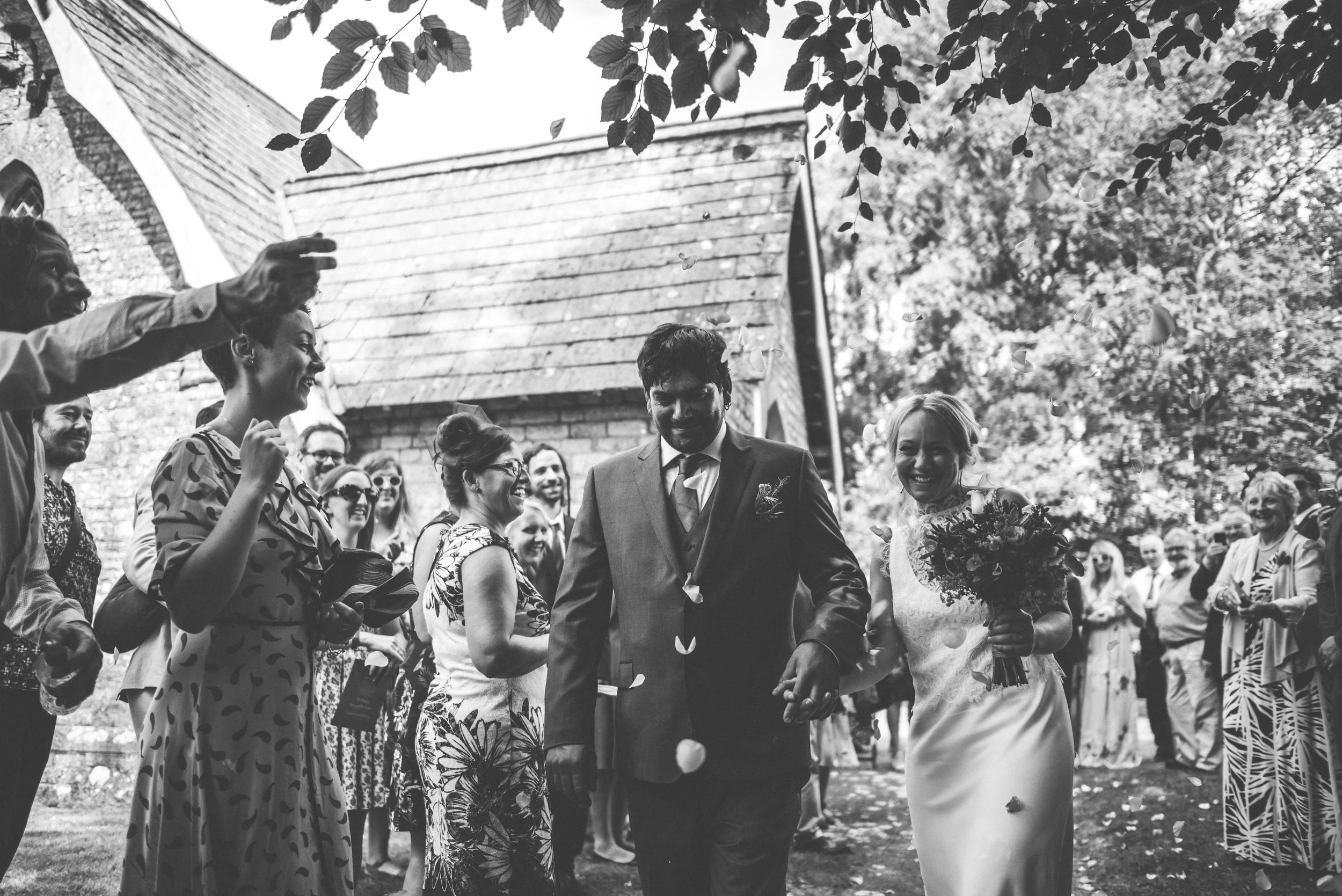 Wedding captured by Cornwall wedding photographer Mark Shaw Photography - A black and white photo of a wedding celebration outdoors, showing a couple walking hand-in-hand through a crowd of smiling guests, with one woman holding a bouquet of flowers.