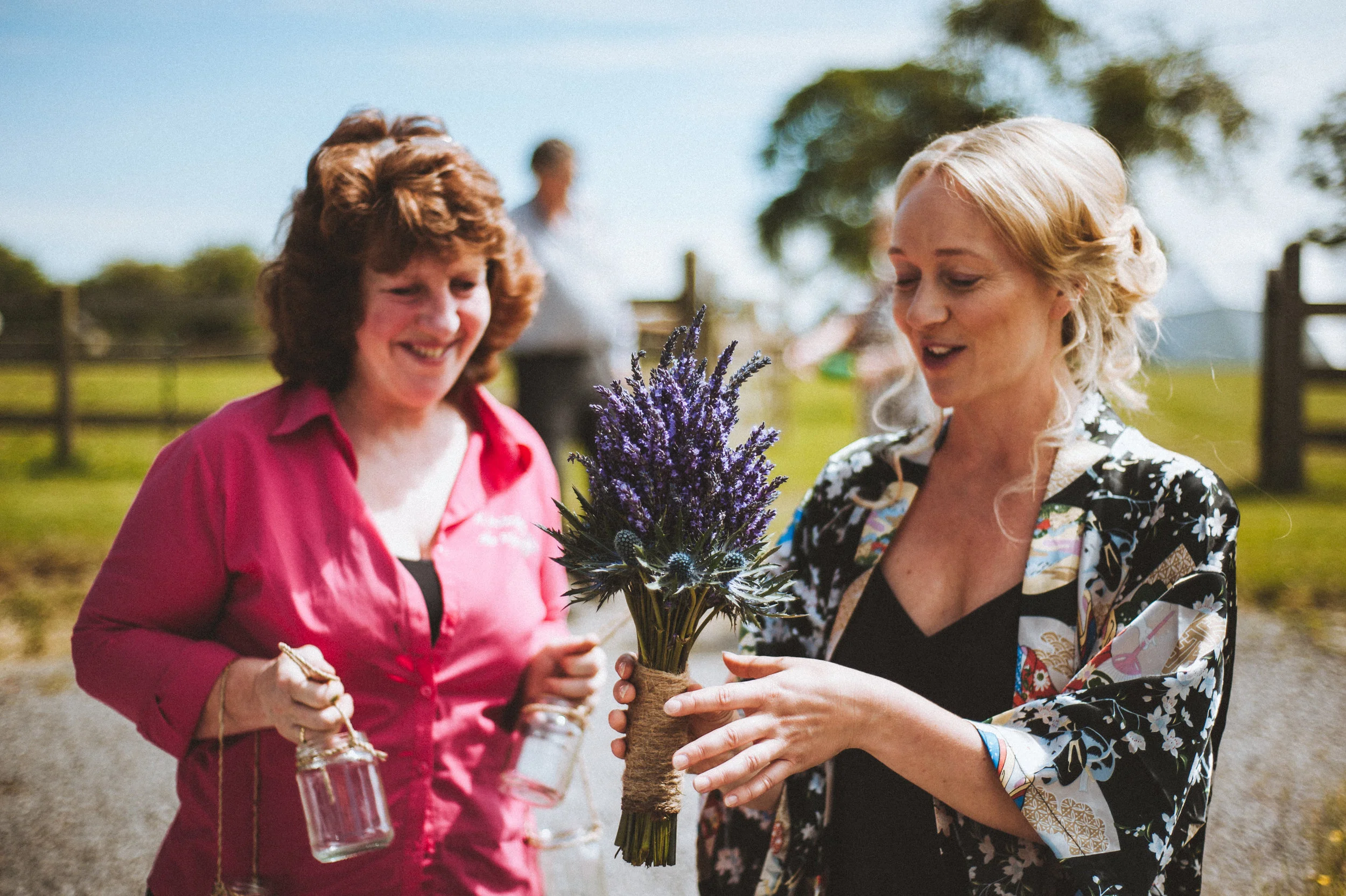 Two women standing outdoors, one holding a small bouquet of purple flowers, smiling and looking at the flowers, with a blurry person and fence in the background.