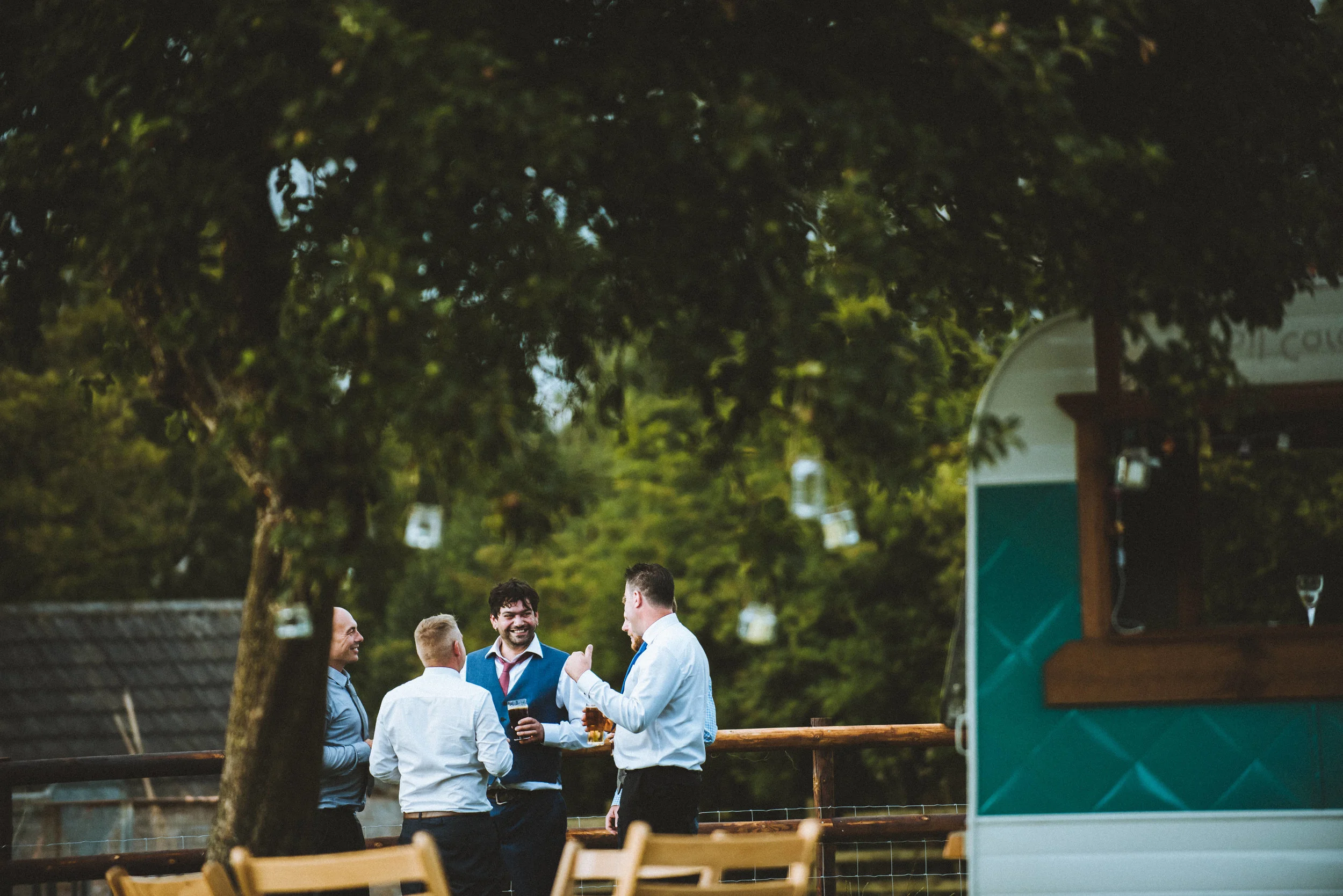 Wedding captured by Cornwall wedding photographer Mark Shaw Photography - Four men dressed in semi-formal attire engaging in conversation while holding drinks, in an outdoor setting with trees and wooden chairs, next to a food truck.