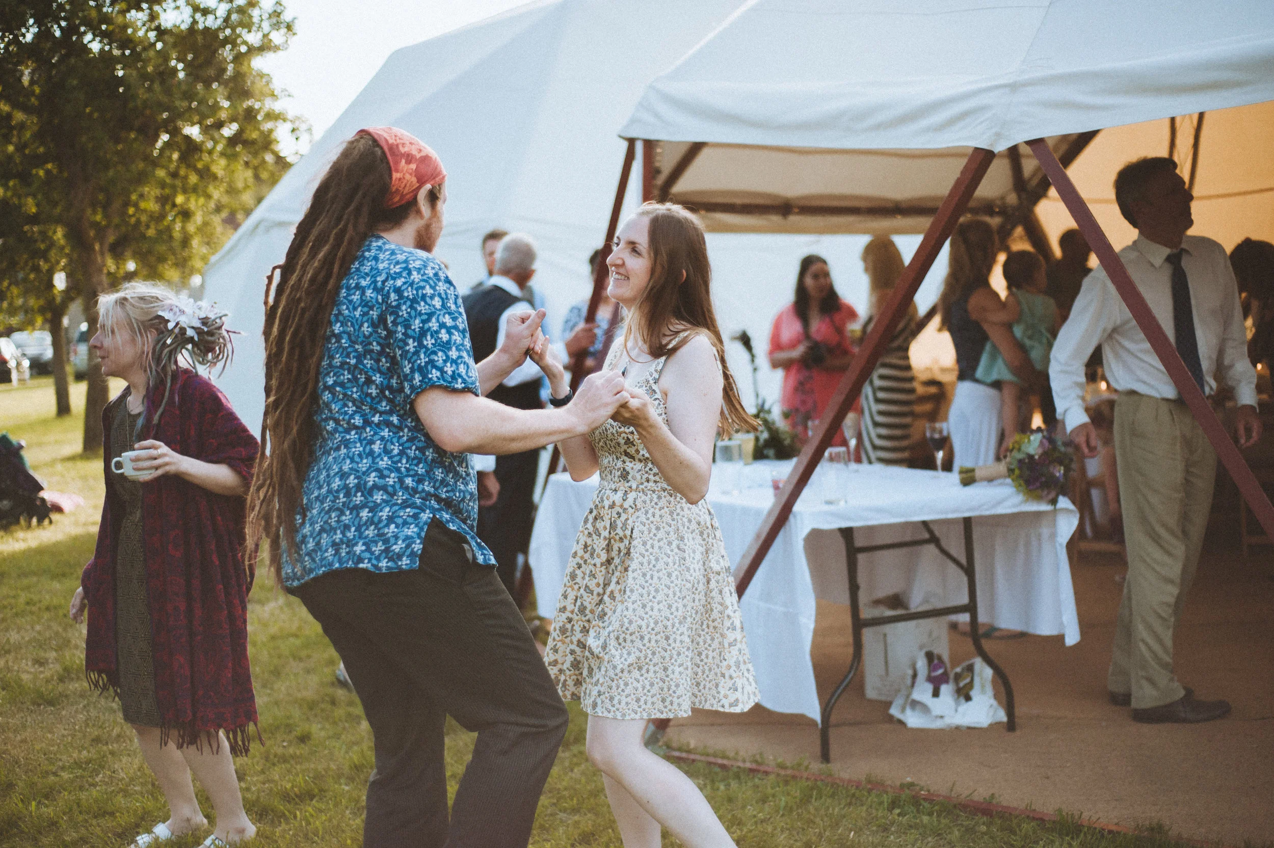 Wedding captured by Cornwall wedding photographer Mark Shaw Photography - People dancing and socializing at an outdoor gathering with a tent and tables in the background, during daylight.