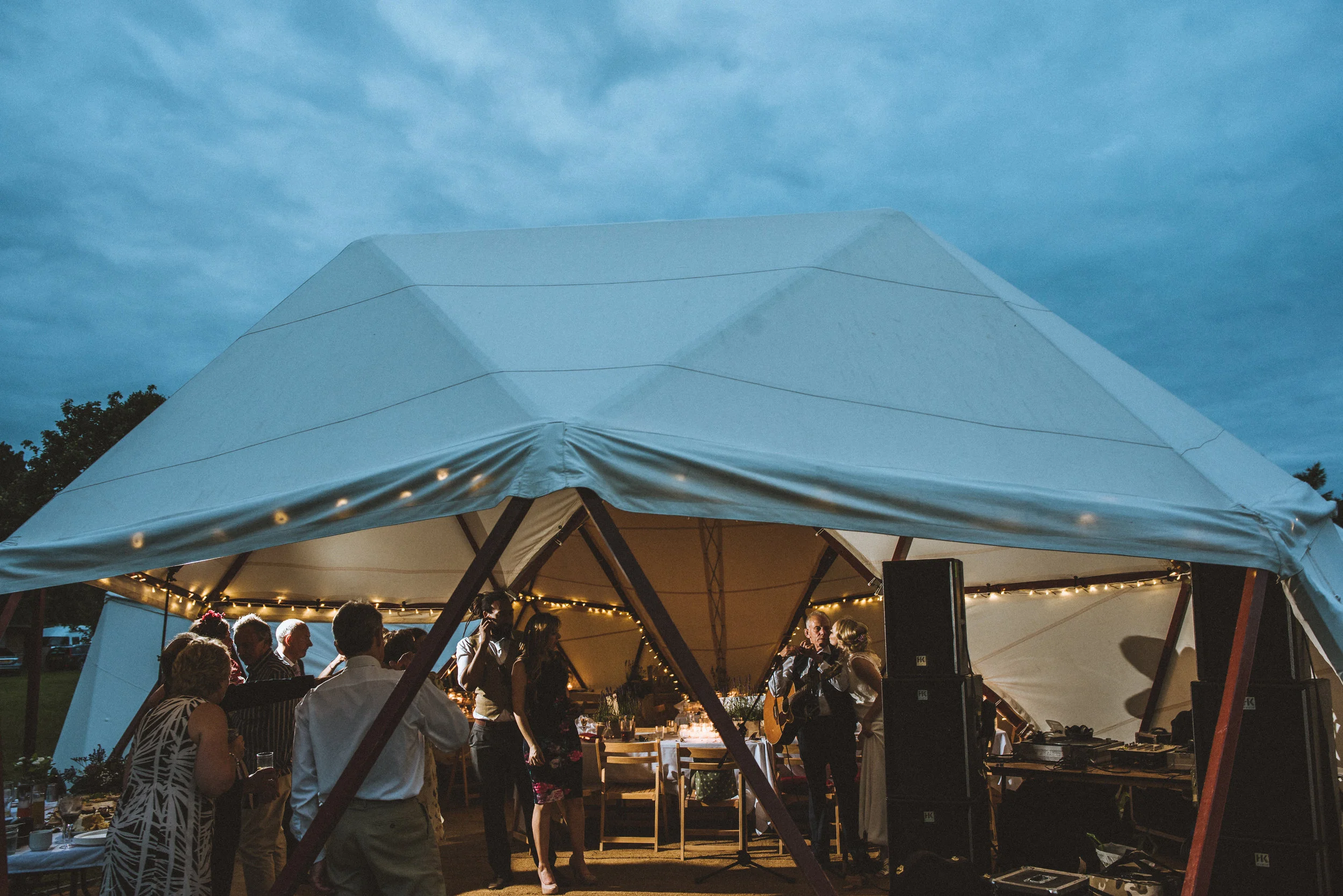 Tipi wedding captured by Cornwall wedding photographer Mark Shaw Photography - Guests dancing and socializing under a large, white, open-sided tent with string lights, at an outdoor evening event.