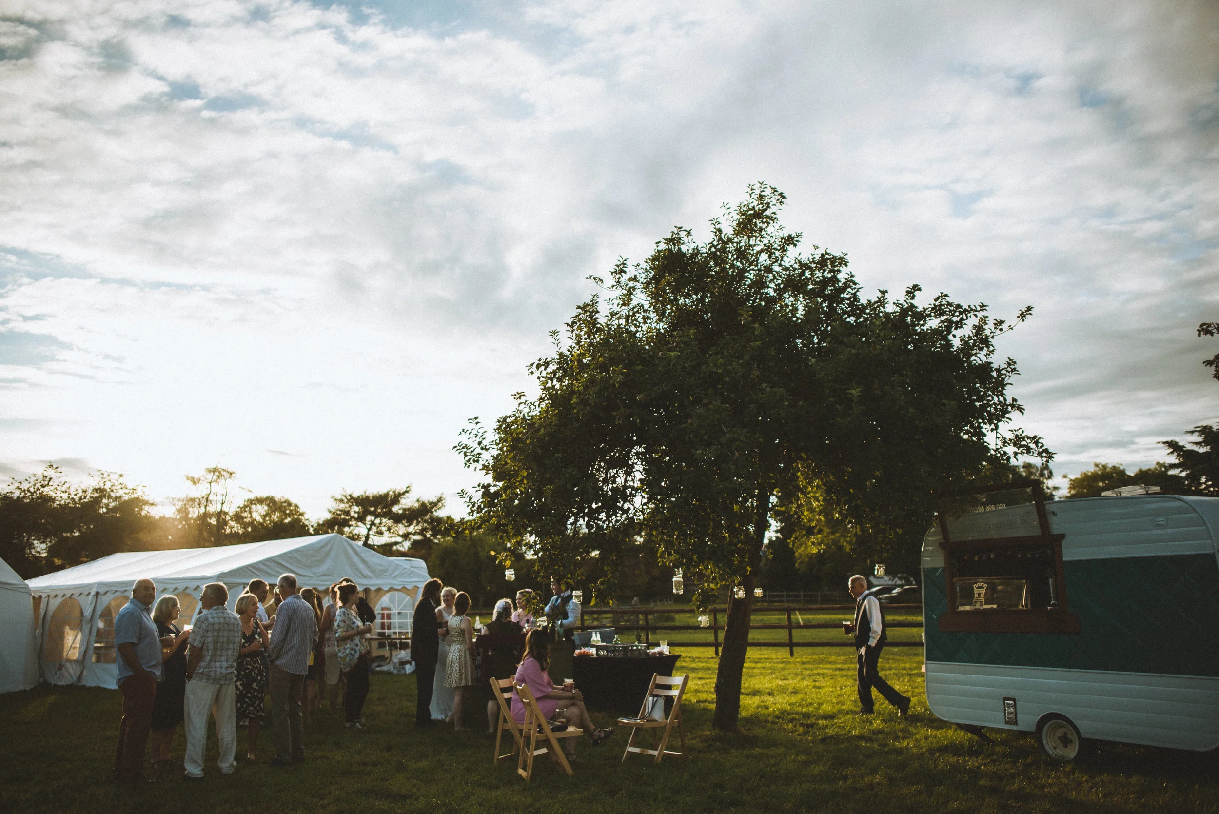 Wedding captured by Cornwall wedding photographer Mark Shaw Photography - People gathering outdoors near a tent and a food truck during a social event at sunset, with a large tree and cloudy sky in the background.
