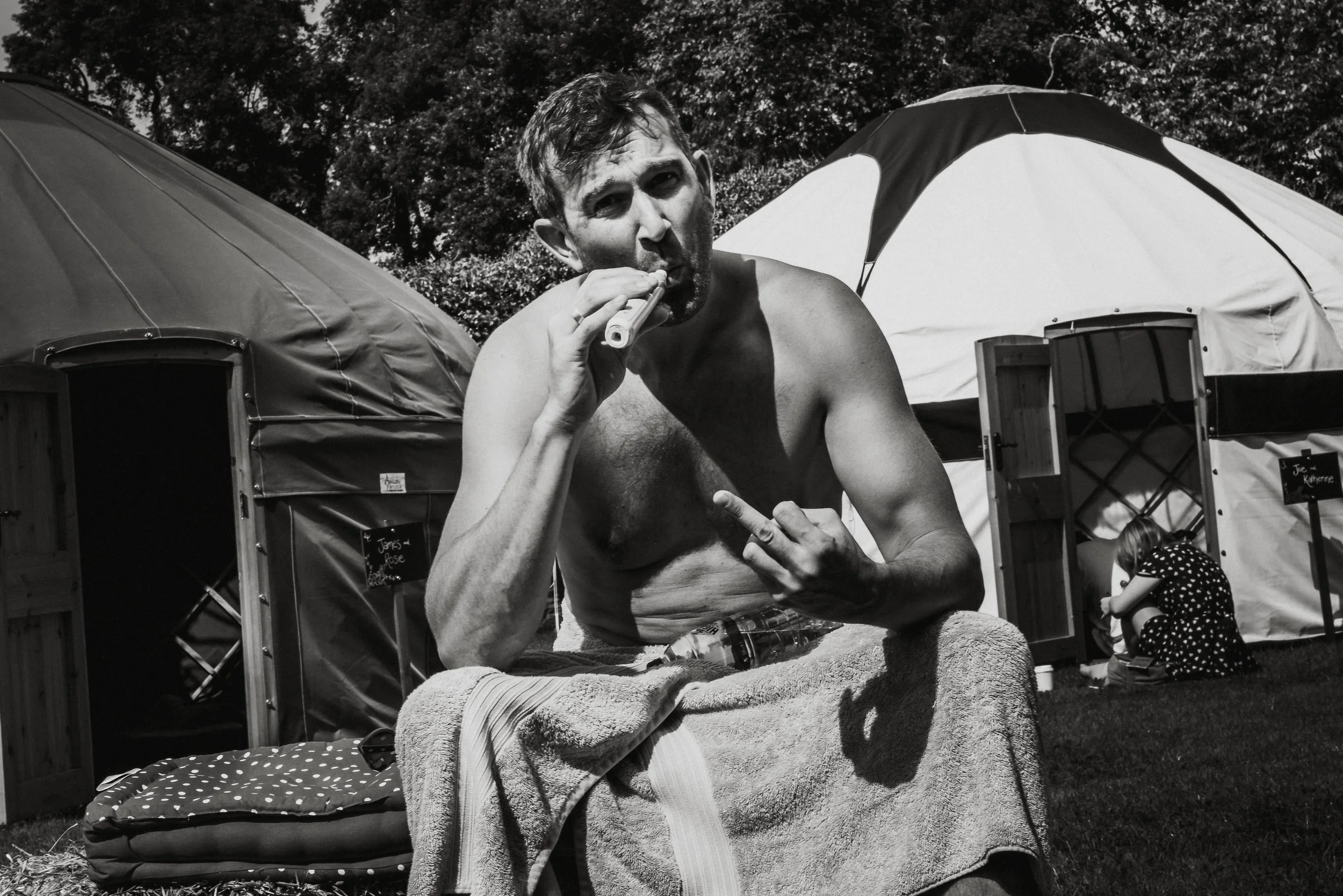 A shirtless man with short hair sitting outdoors, holding a vape in one hand and making a gesture with the other hand. Behind him are two tents and a girl sitting on the grass, wearing a polka-dot dress, and appearing to work on something near one te