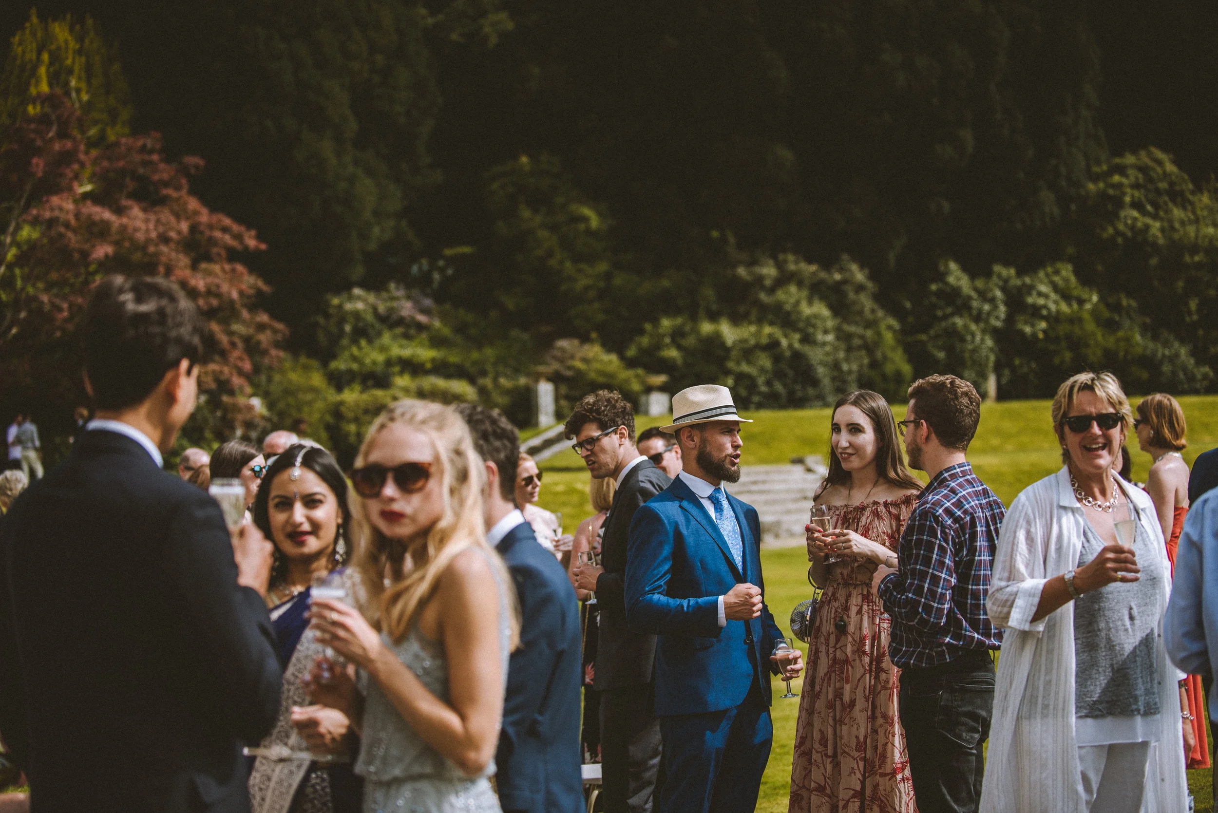 People at an outdoor social event in a park, dressed in formal and semi-formal clothing, holding drinks and engaging in conversations amid green trees and grass.