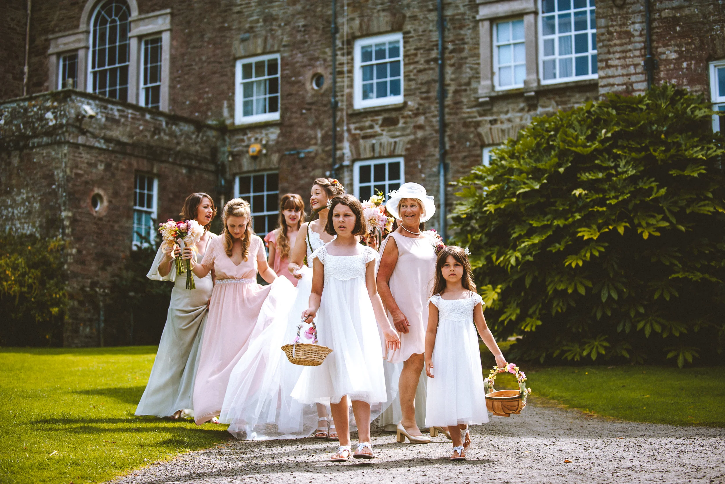 Group of women and girls in white and pastel dresses walking on a gravel path outside a historic brick building, holding flowers and baskets, during a wedding or special event.