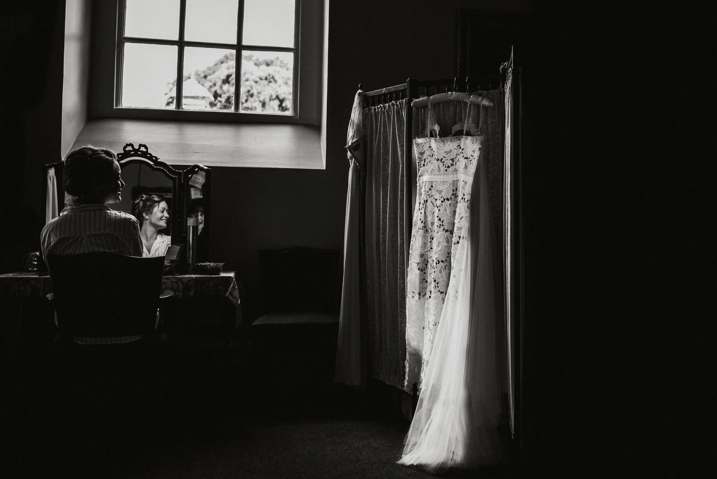 A woman sitting at a vanity table with a mirror, smiling as she looks at herself, with her reflection visible. In the foreground, a clothing rack displays lace and chiffon wedding dresses, illuminated by natural light from a window.