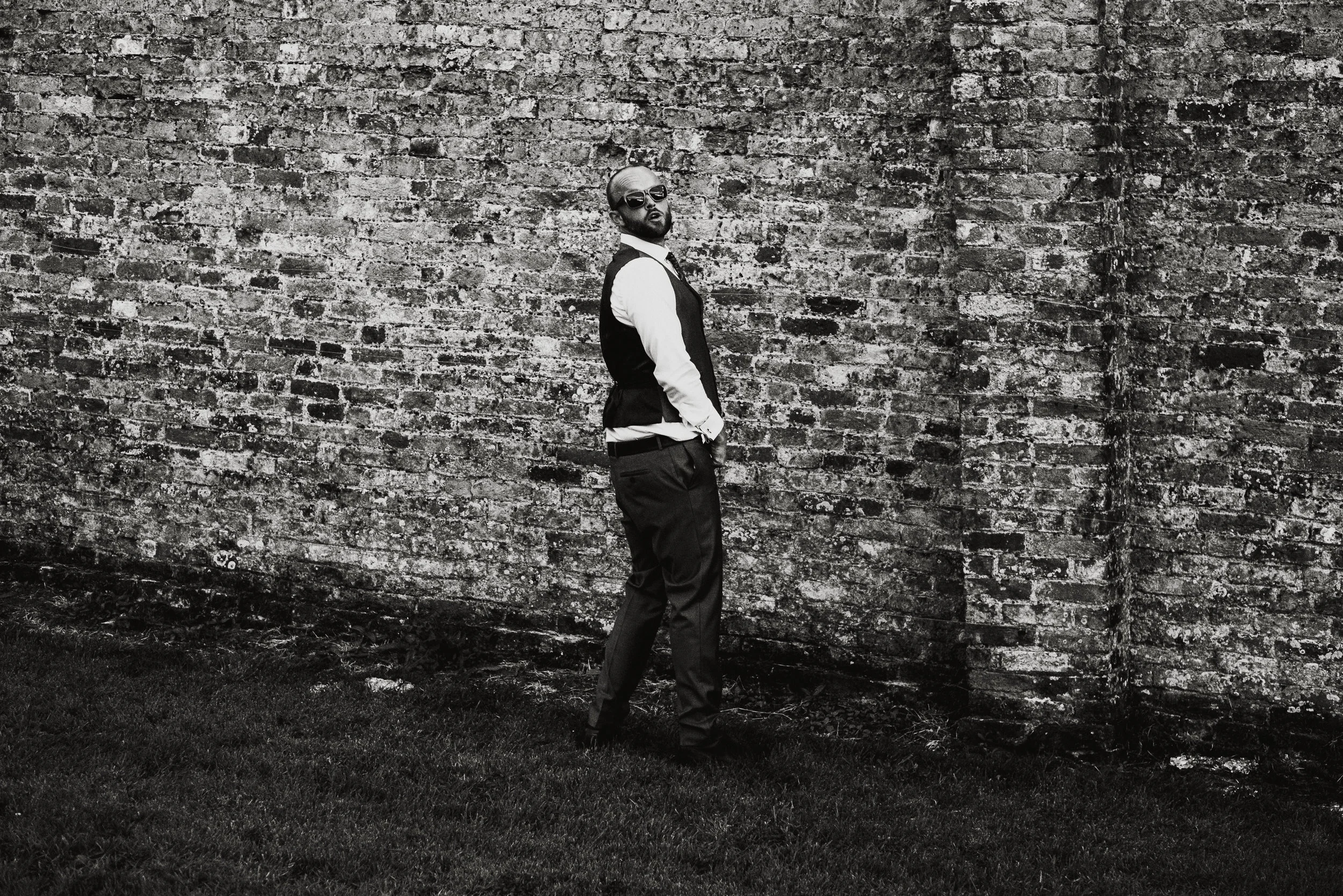 Man wearing sunglasses, white shirt, and dark vest standing against a brick wall, looking towards the camera.