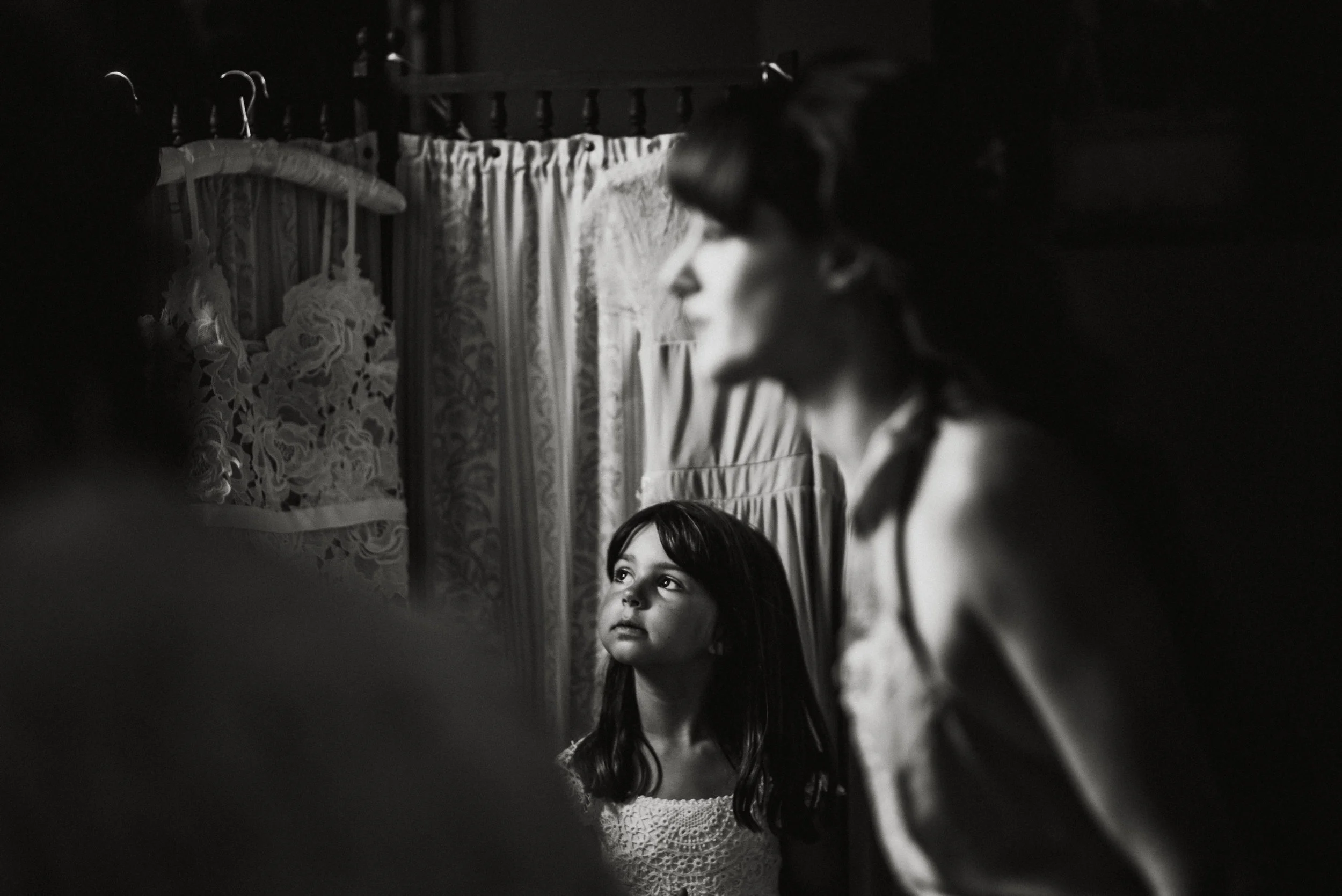 A young girl is looking upward with a serious expression. A woman, partially out of focus, stands nearby in a room with patterned curtains and floral decorations. The photo is in black and white.