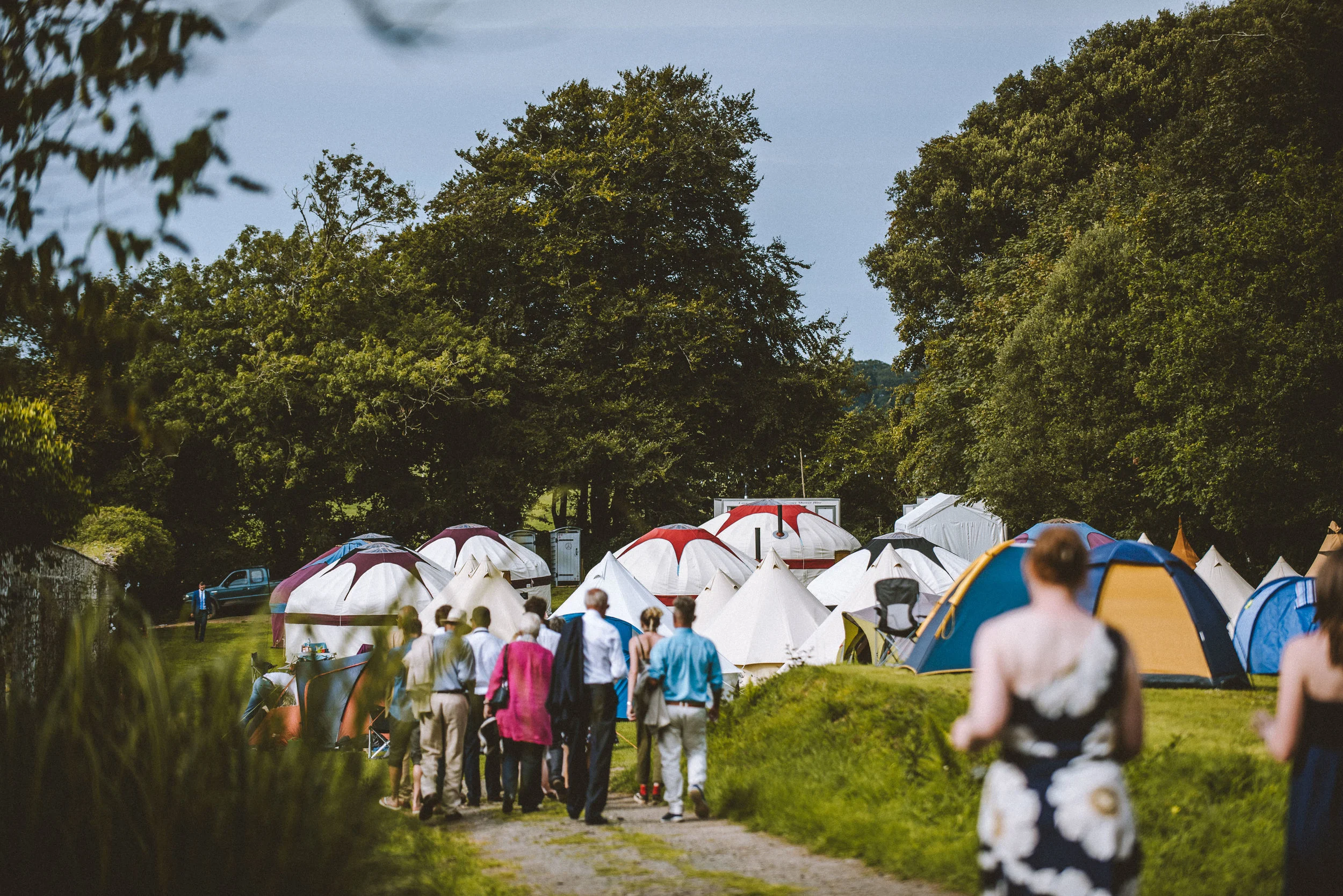 People walking on a path towards a campsite with numerous tents set up among trees.