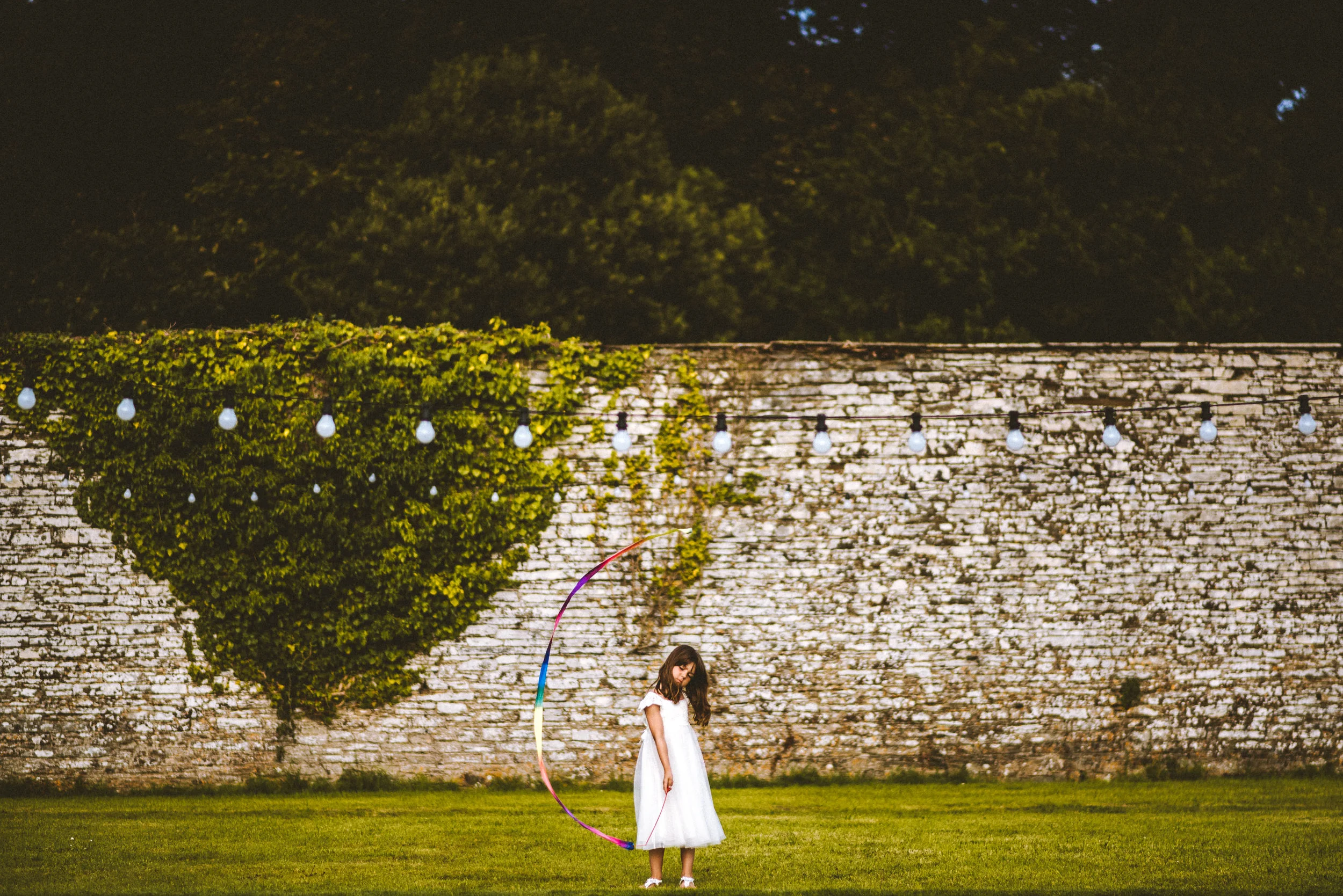 A young girl in a white dress stands on a grassy area at night, holding a large rainbow-colored hula hoop, with a stone wall and string lights in the background.