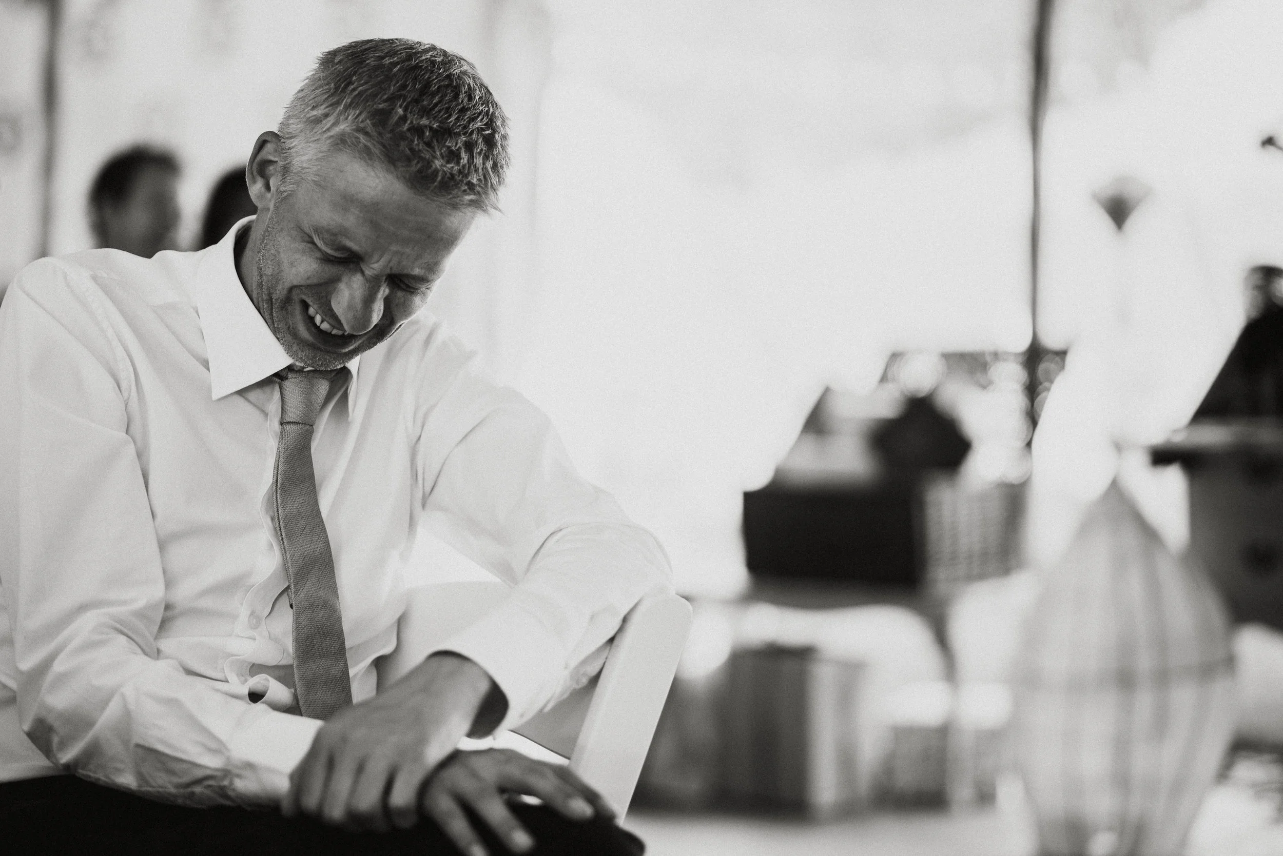 A man in a white shirt and tie smiling and leaning forward in an office or waiting area, with blurred people and furniture in the background.
