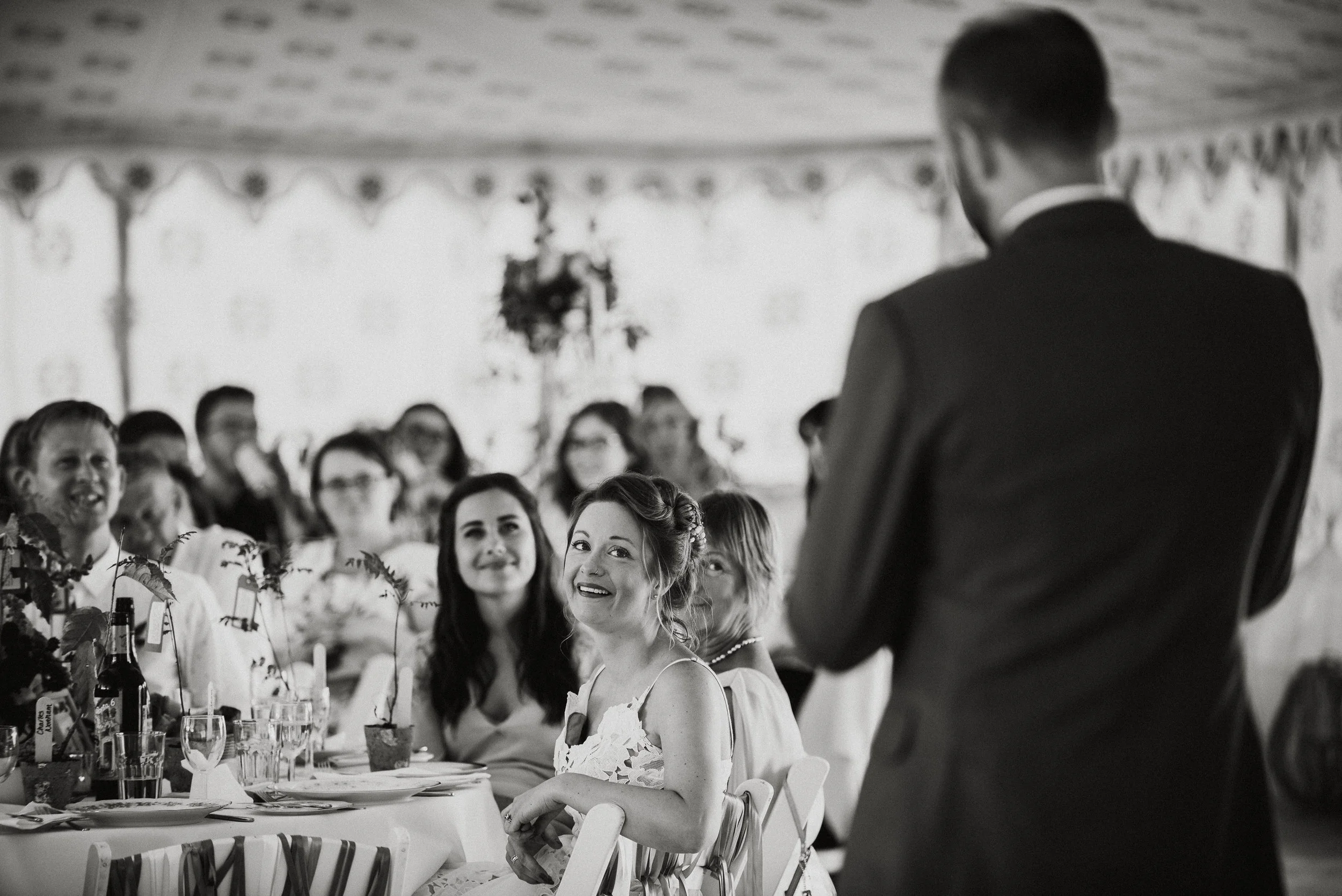 A woman at a wedding reception smiles while looking at a man who appears to be giving a speech. She sits at a decorated table with other guests in the background.