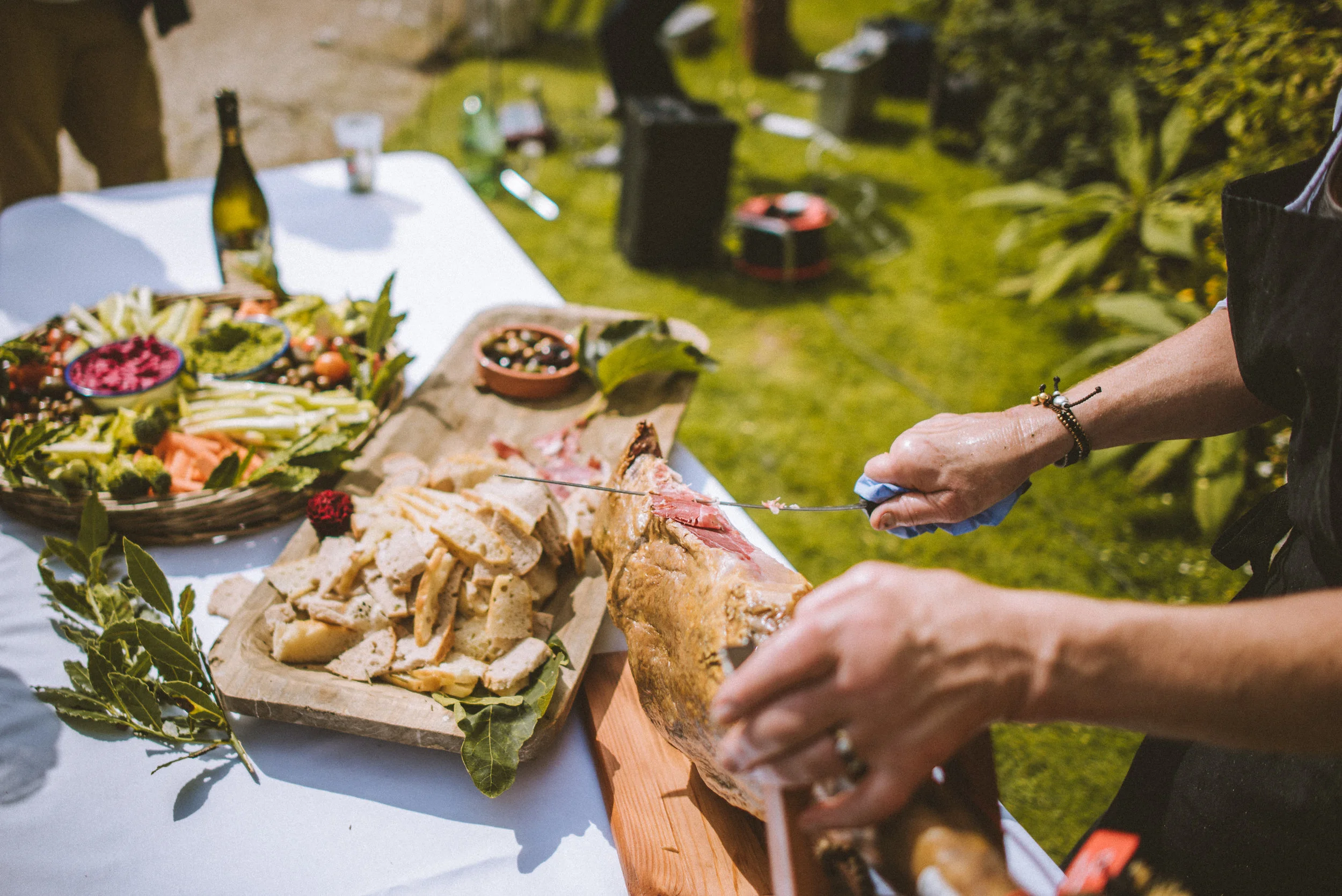 Person slicing a ham at an outdoor picnic table with assorted food including bread, vegetables, and a salad.