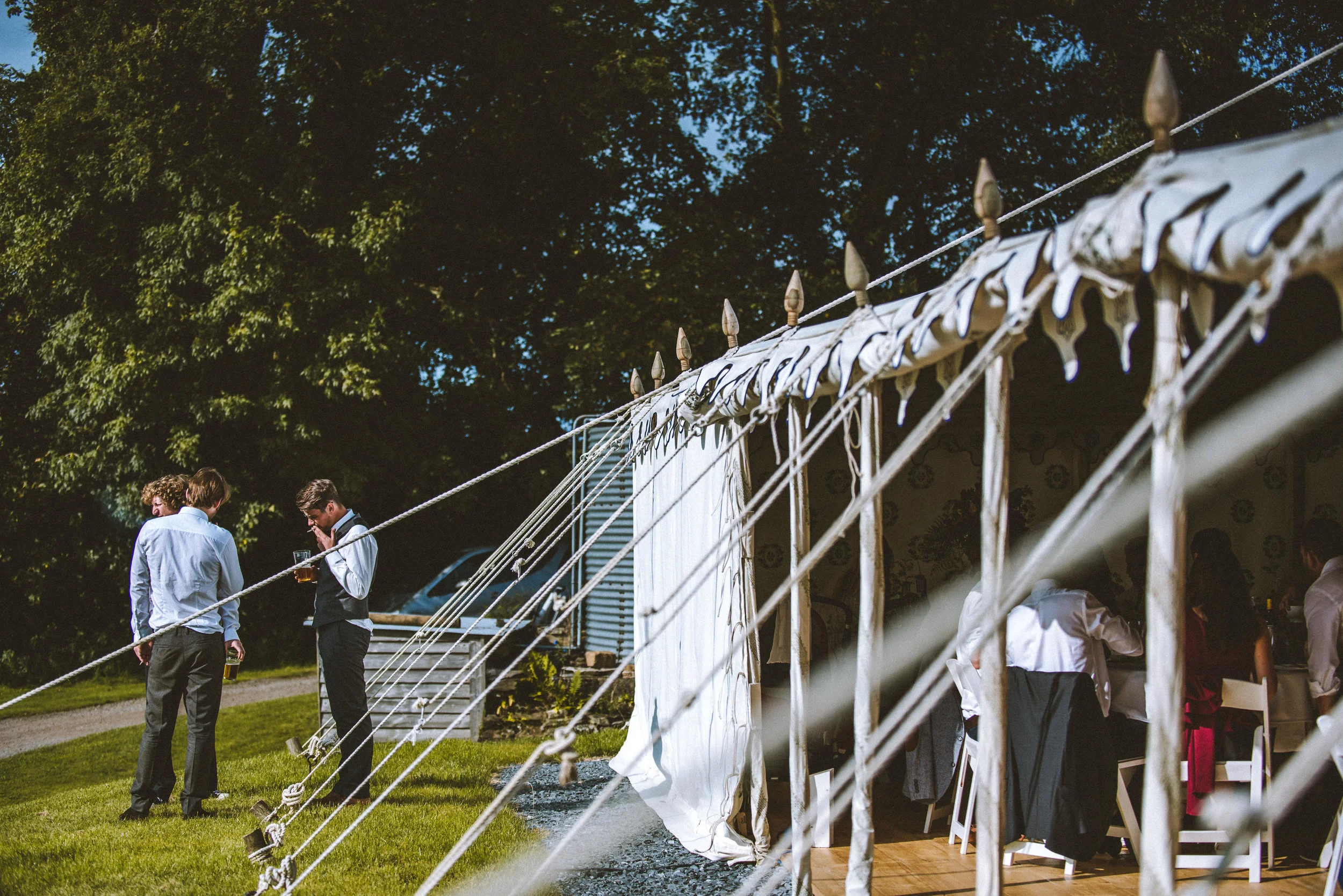 People dressed formally standing outside a large white event tent with open sides, set on a grassy area surrounded by trees.