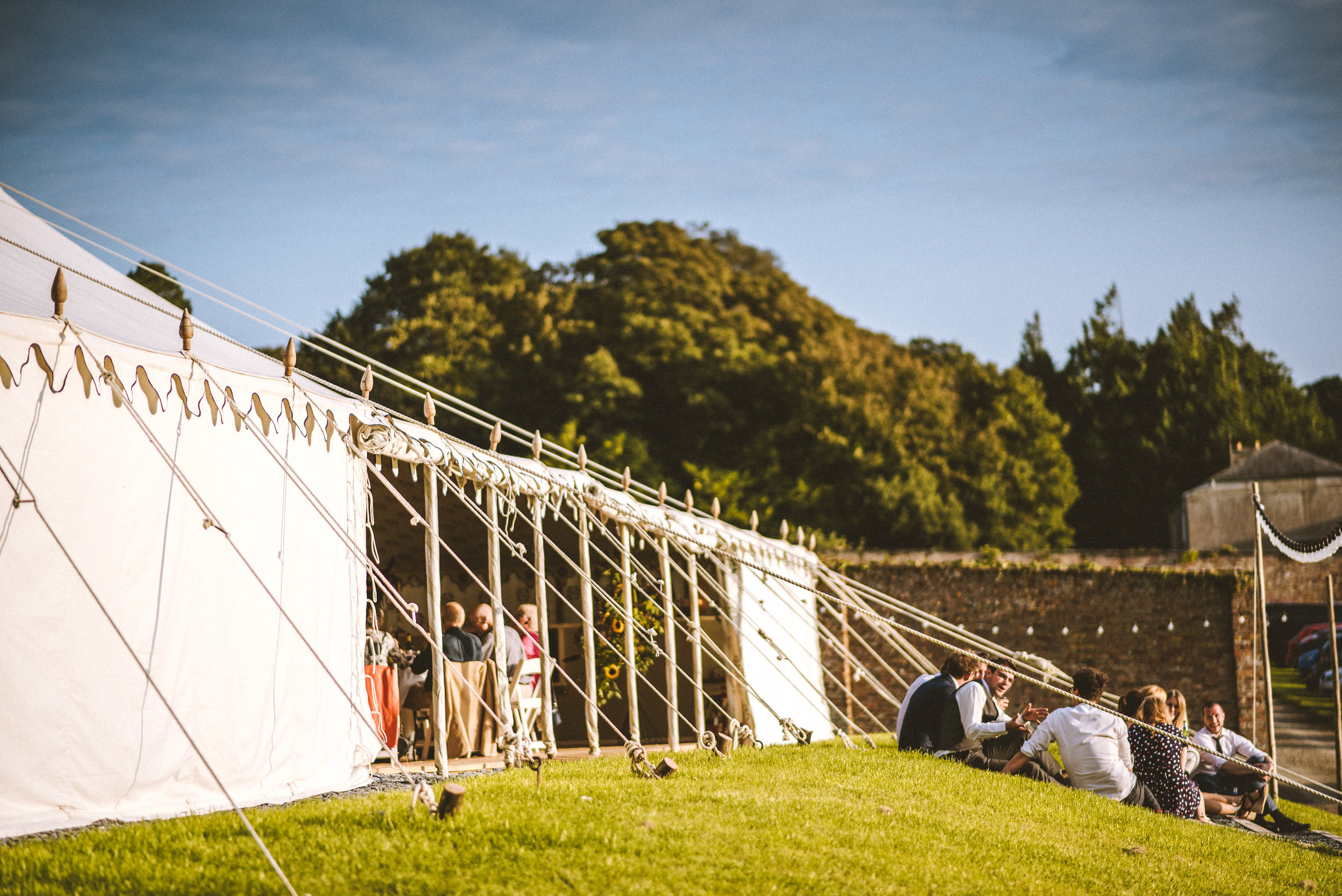 A large outdoor event tent with people sitting under it and on the grass outside on a sunny day, with trees and a hill in the background.
