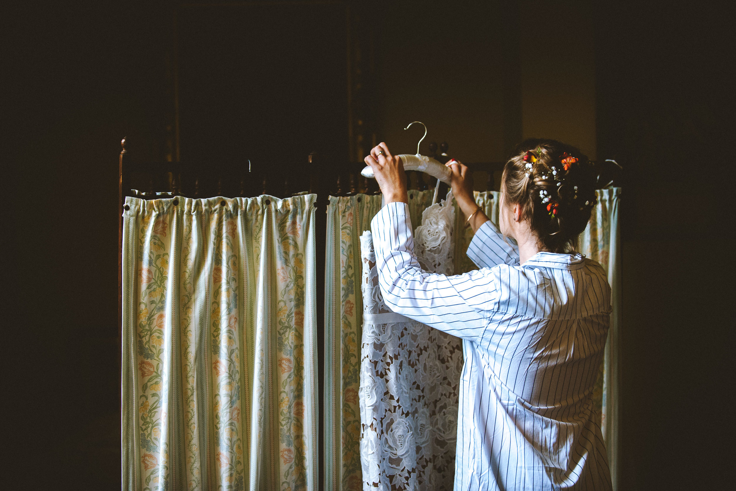A woman with floral hair accessories, wearing a striped shirt, is holding a lace dress on a hanger in front of a fabric divider.