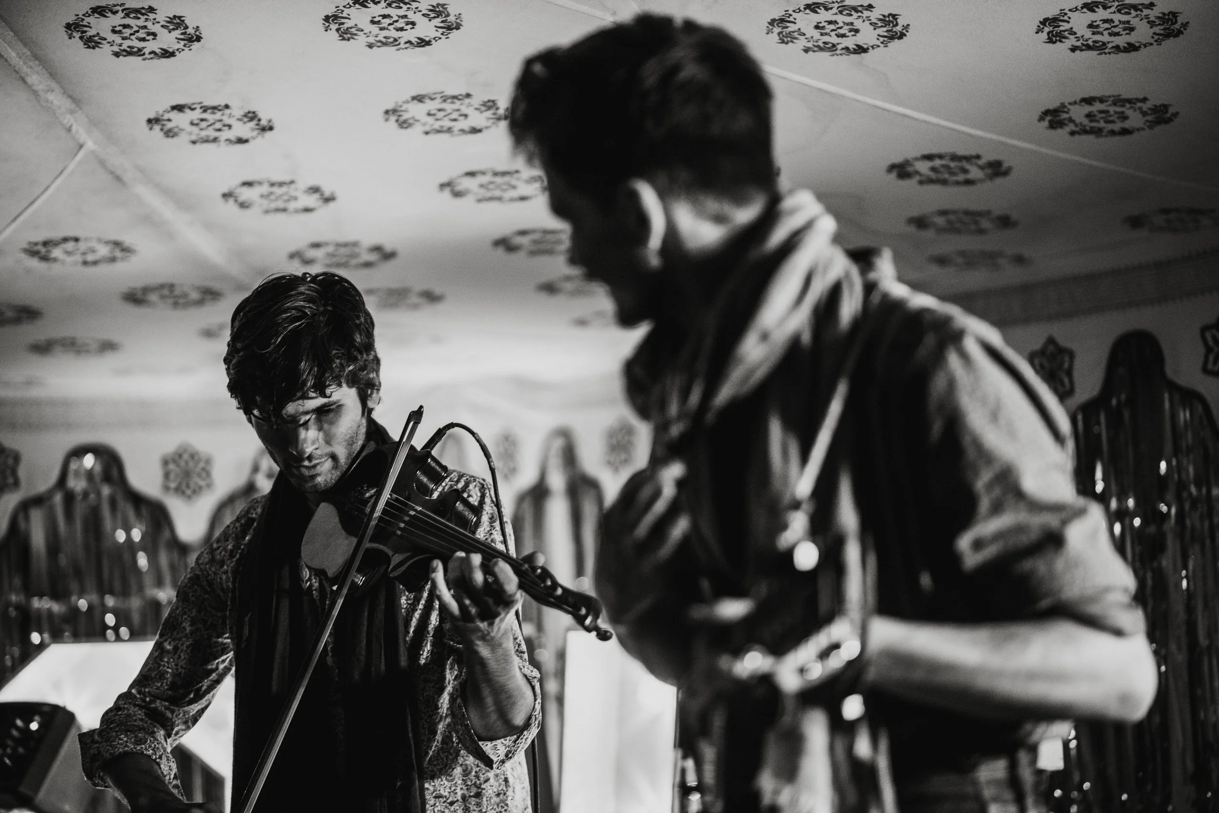 Two men playing string instruments, one on a violin and the other on a guitar, in a room with ornate decorations on the ceiling.