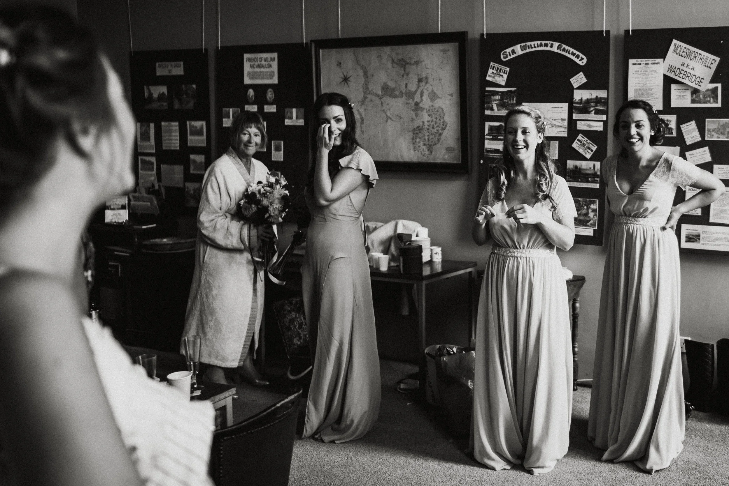Black and white photo of a bridal party, with four women in dresses standing in a room decorated with posters and a large map. One is holding a bouquet, and some are smiling or laughing.