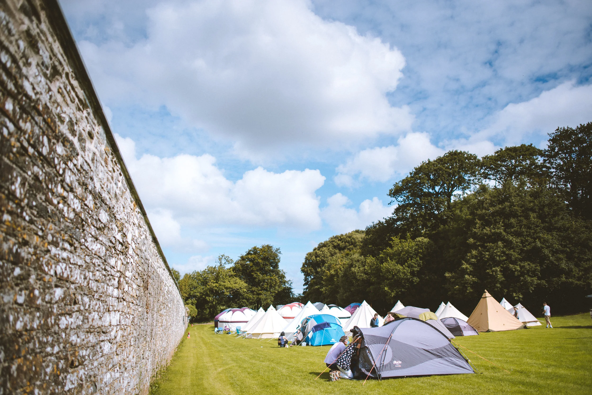 A tent camping site on a grassy field with multiple tents and people, a stone wall on the left, trees in the background, and blue sky with clouds overhead.
