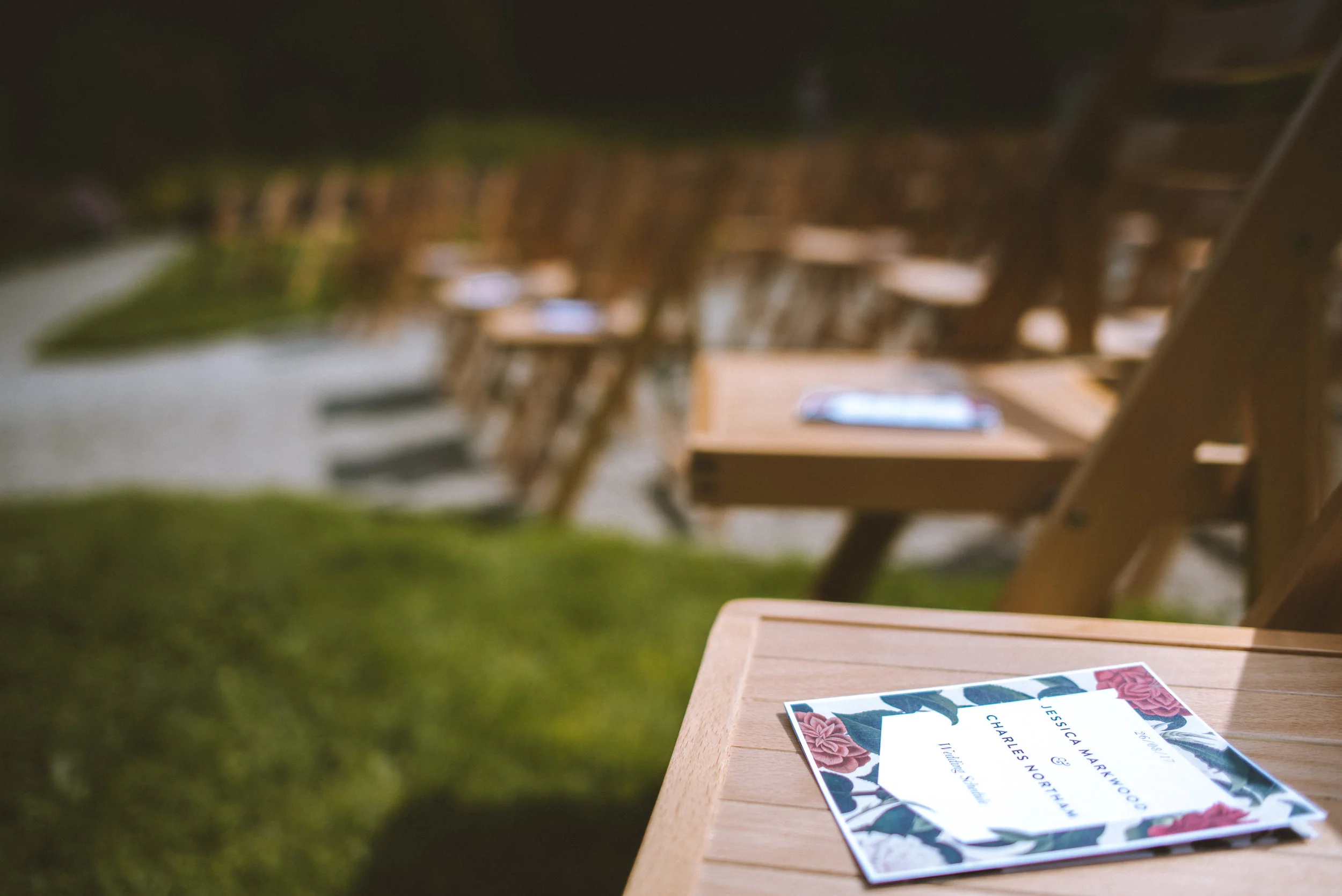 Wedding invitation on a wooden table at an outdoor event with empty chairs in the background.