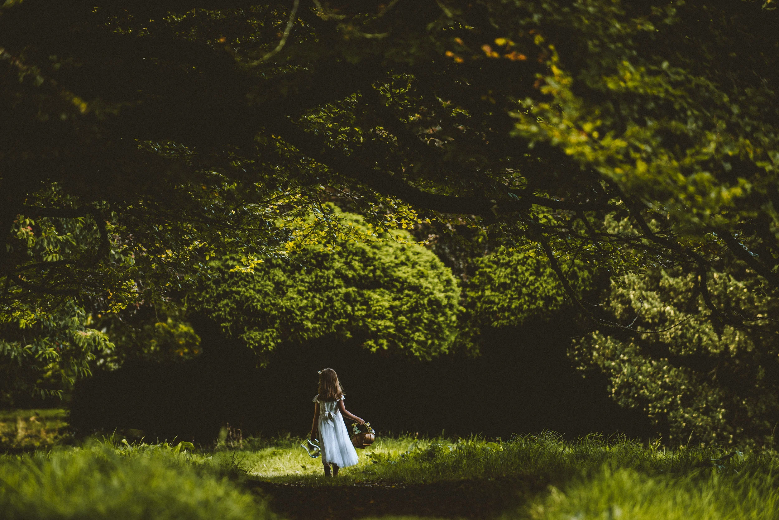 A young girl in a white dress walking along a shaded forest trail, carrying a basket and a pair of shoes.