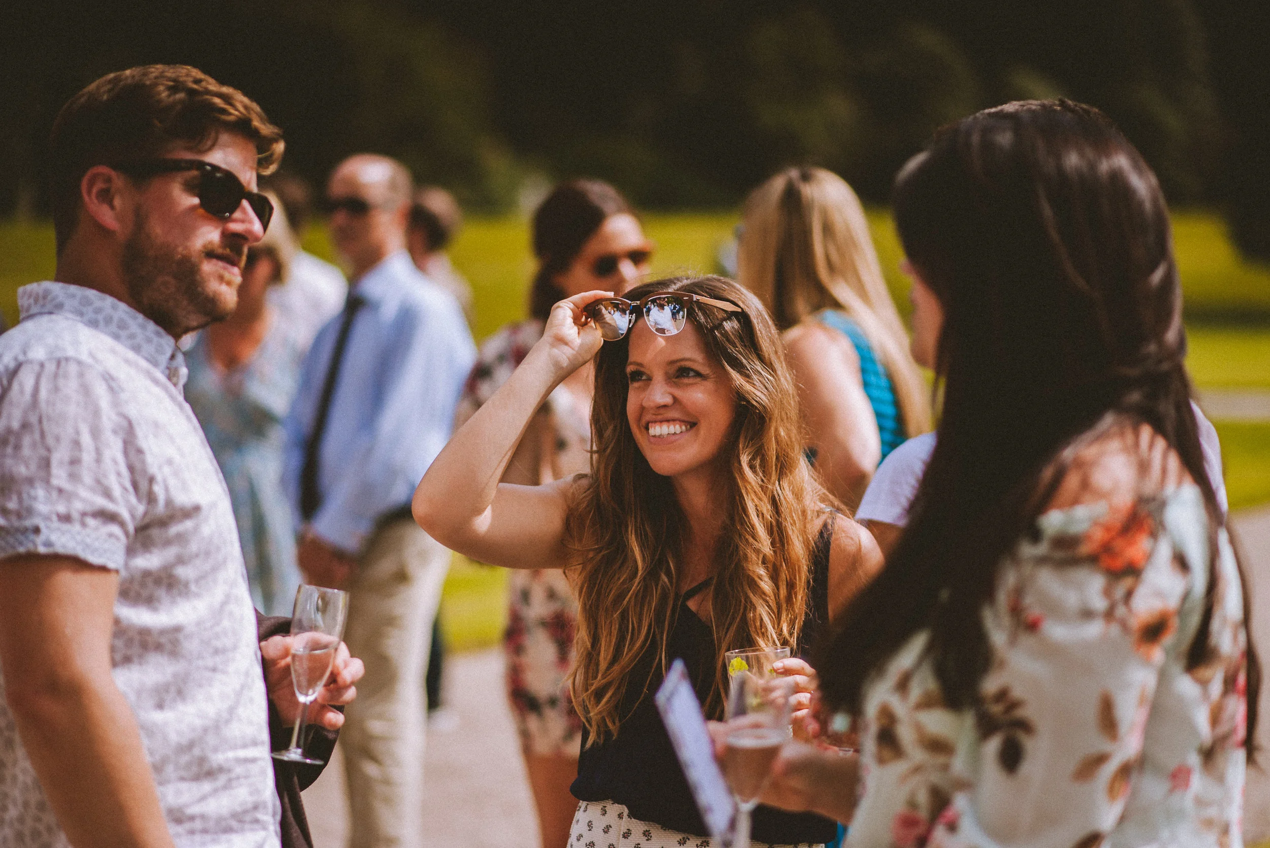 A group of people socializing outdoors during daytime, smiling, holding drinks, with a woman with long hair wearing sunglasses on her head in the center.