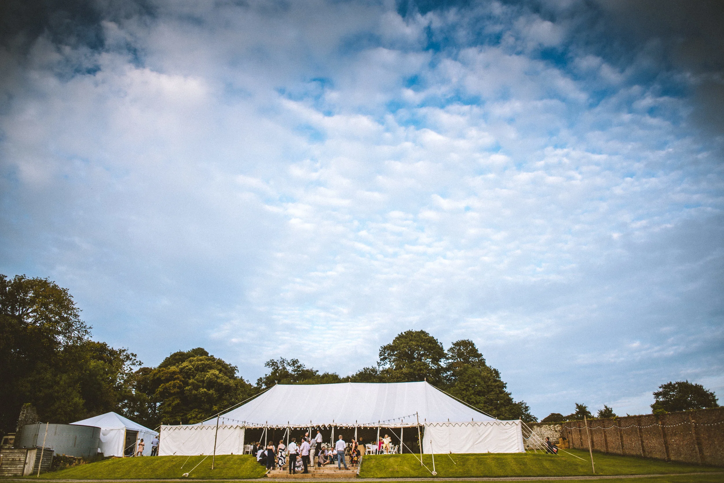 Large white event tent set up outdoors on green grass with people gathered under the tent, trees in the background, and a cloudy sky overhead.