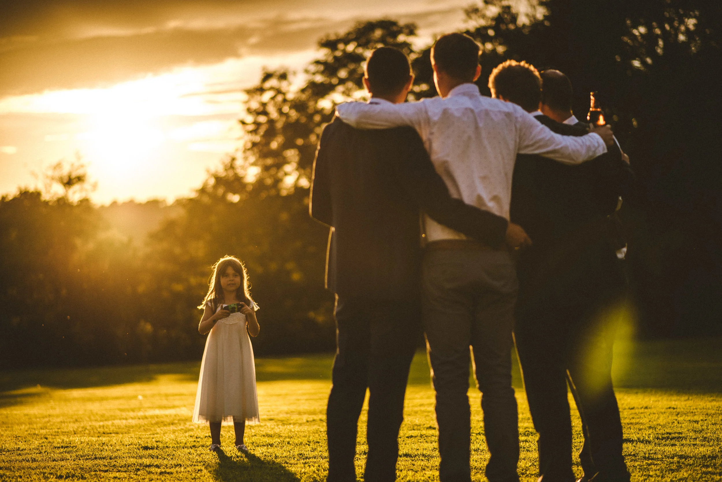 A group of young men with arms around each other, standing on a grassy field at sunset, with a young girl in a white dress holding a camera in the background.
