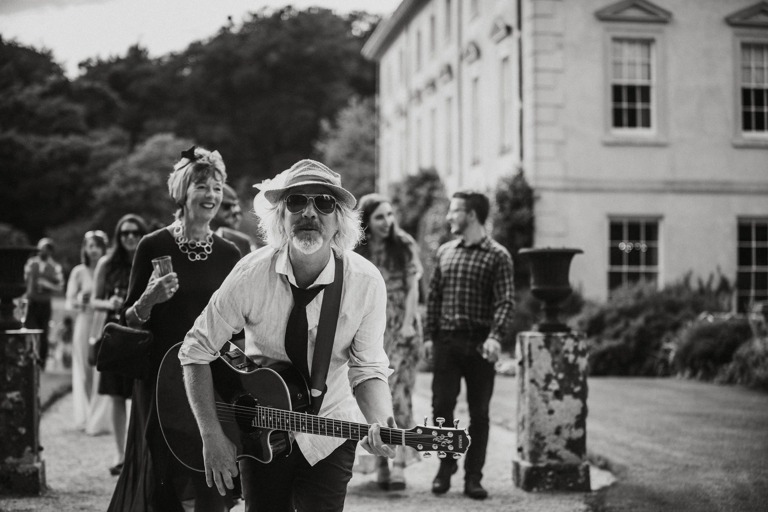 A man playing guitar and singing with a group of people outdoors at a gathering, in front of a large building.
