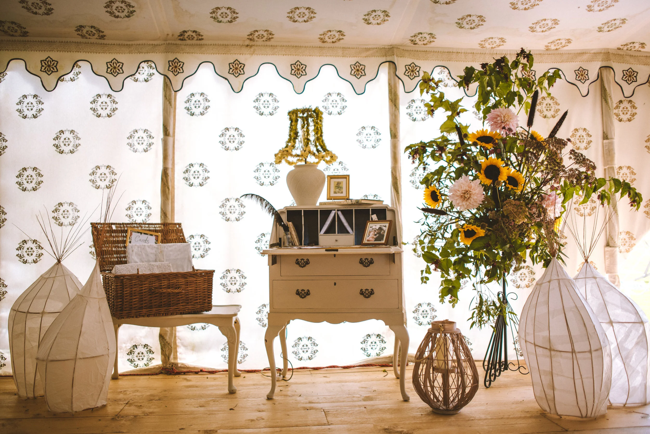 A vintage white desk with a small drawer and ornate handles, against a backdrop of white curtains with floral patterns, topped with a white vase and framed photos. On the left, a wicker basket filled with cards and a white cloth, and two white paper 