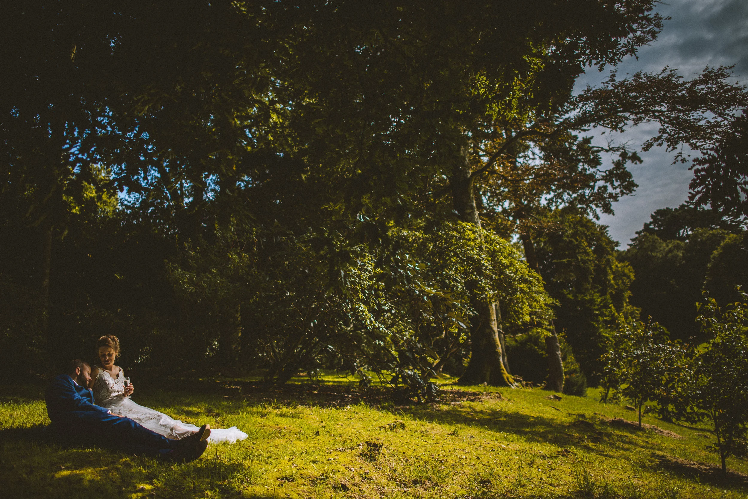A bride and groom sitting on the grass in a wooded area during sunset, surrounded by lush green trees and a partly cloudy sky.