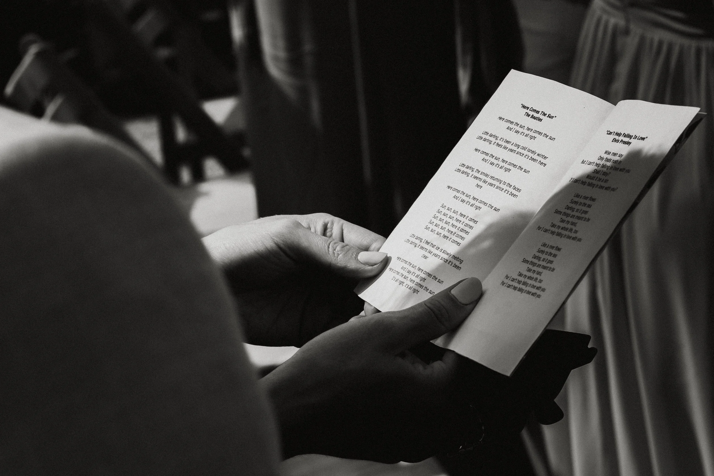 Person holding a piece of paper with printed lyrics, in black and white, with chairs and a table in the background.