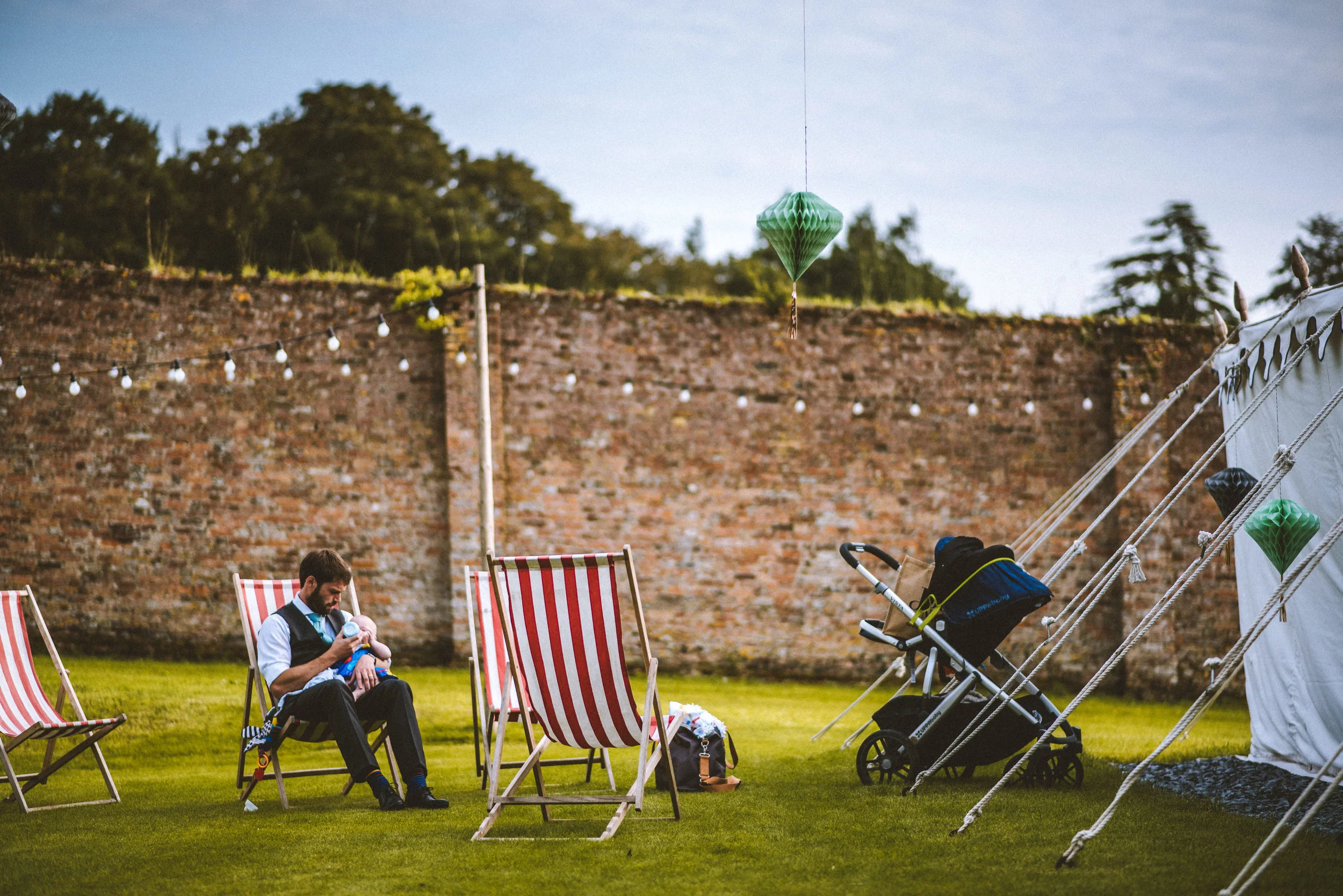 A man sitting on a striped deck chair on a grassy lawn, holding a baby, with another empty striped deck chair nearby. There are decorated lanterns hanging above, a stroller to the right, and a white tent or structure on the right side of the image. A