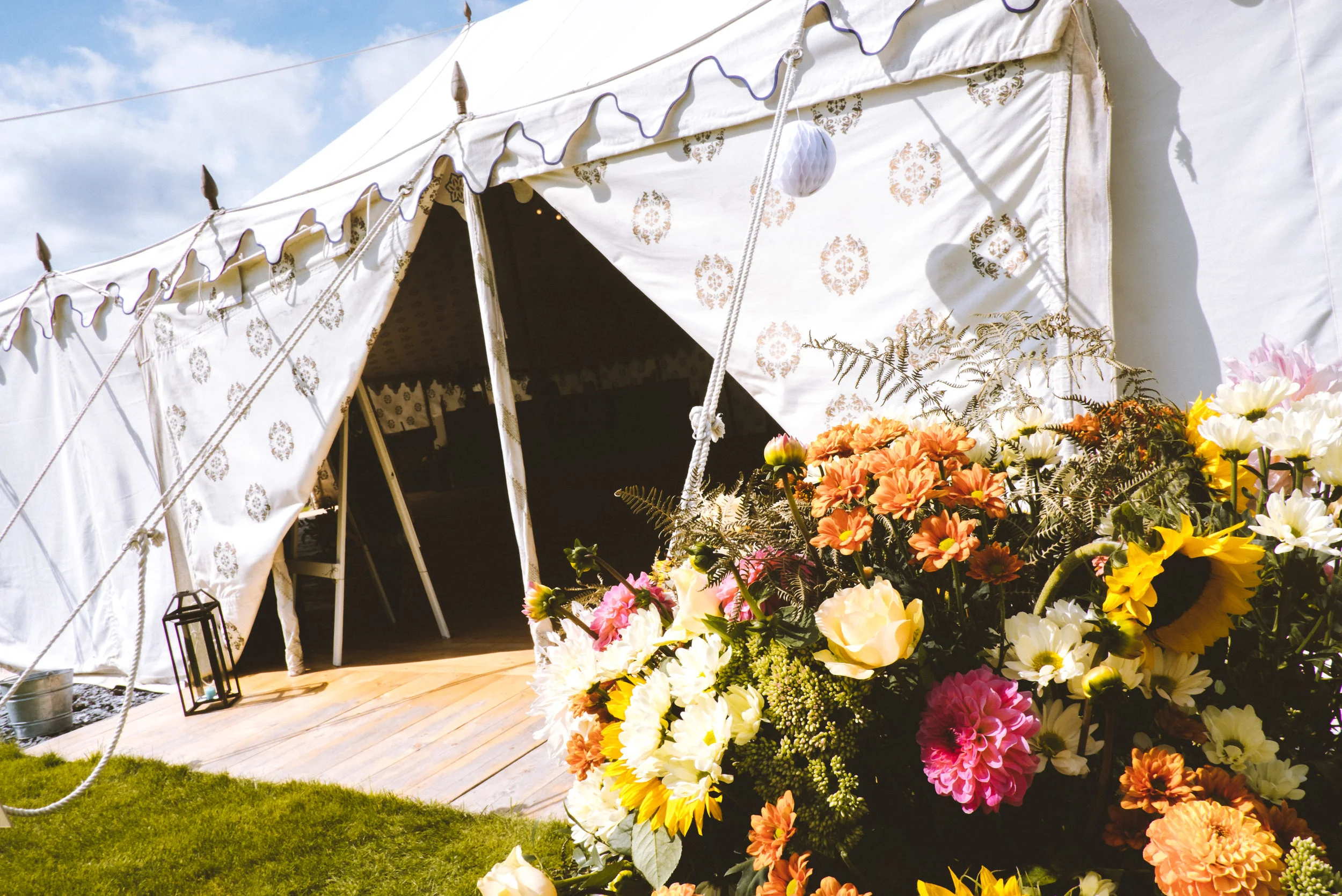 A white tent decorated with patterned fabric, open to reveal a dark interior, with a bouquet of multicolored flowers in the foreground, set on a grassy area under a partly cloudy sky.