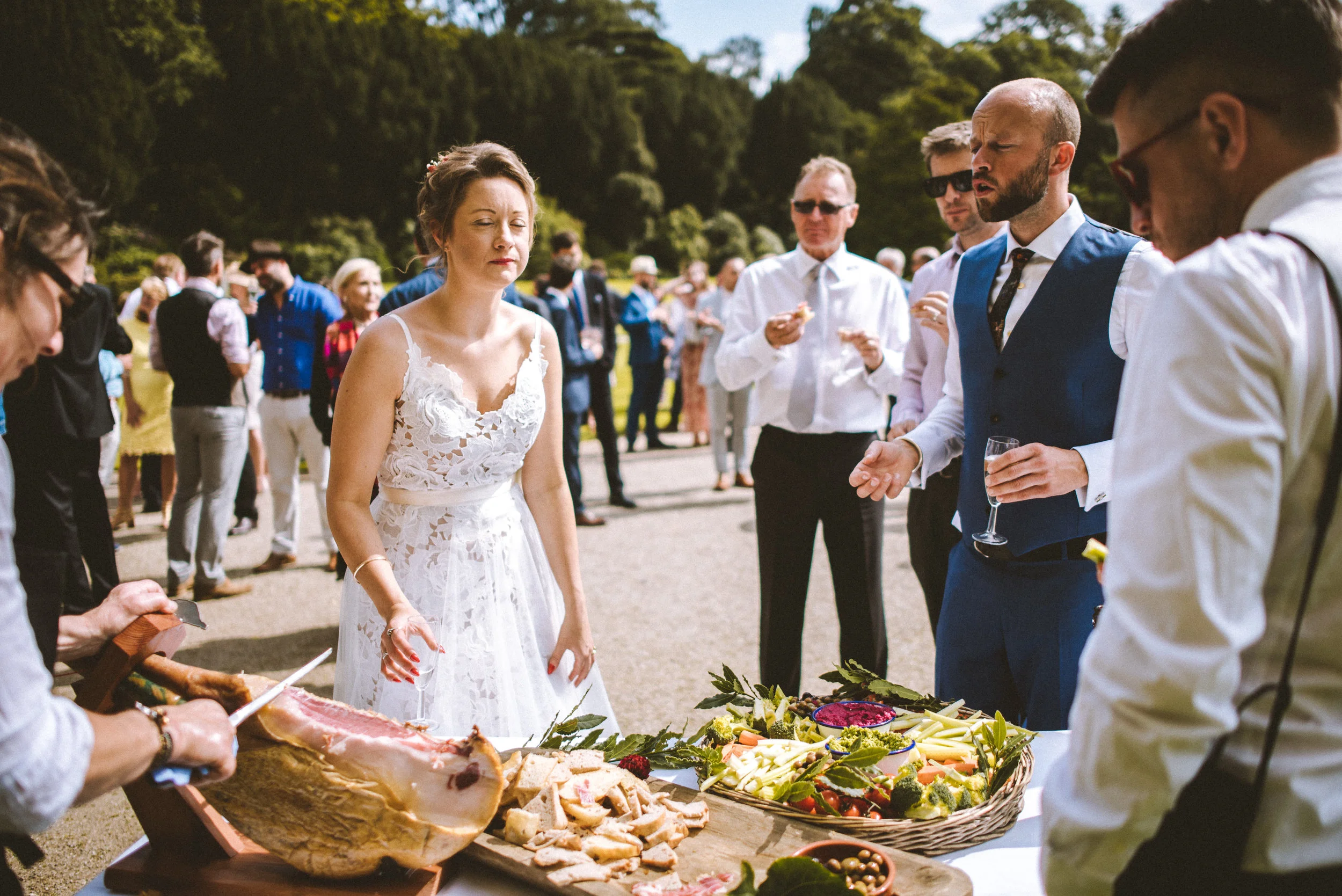 A woman in a white lace dress standing near a food table with various dishes, outdoors at a gathering with several people in formal attire, some holding glasses.